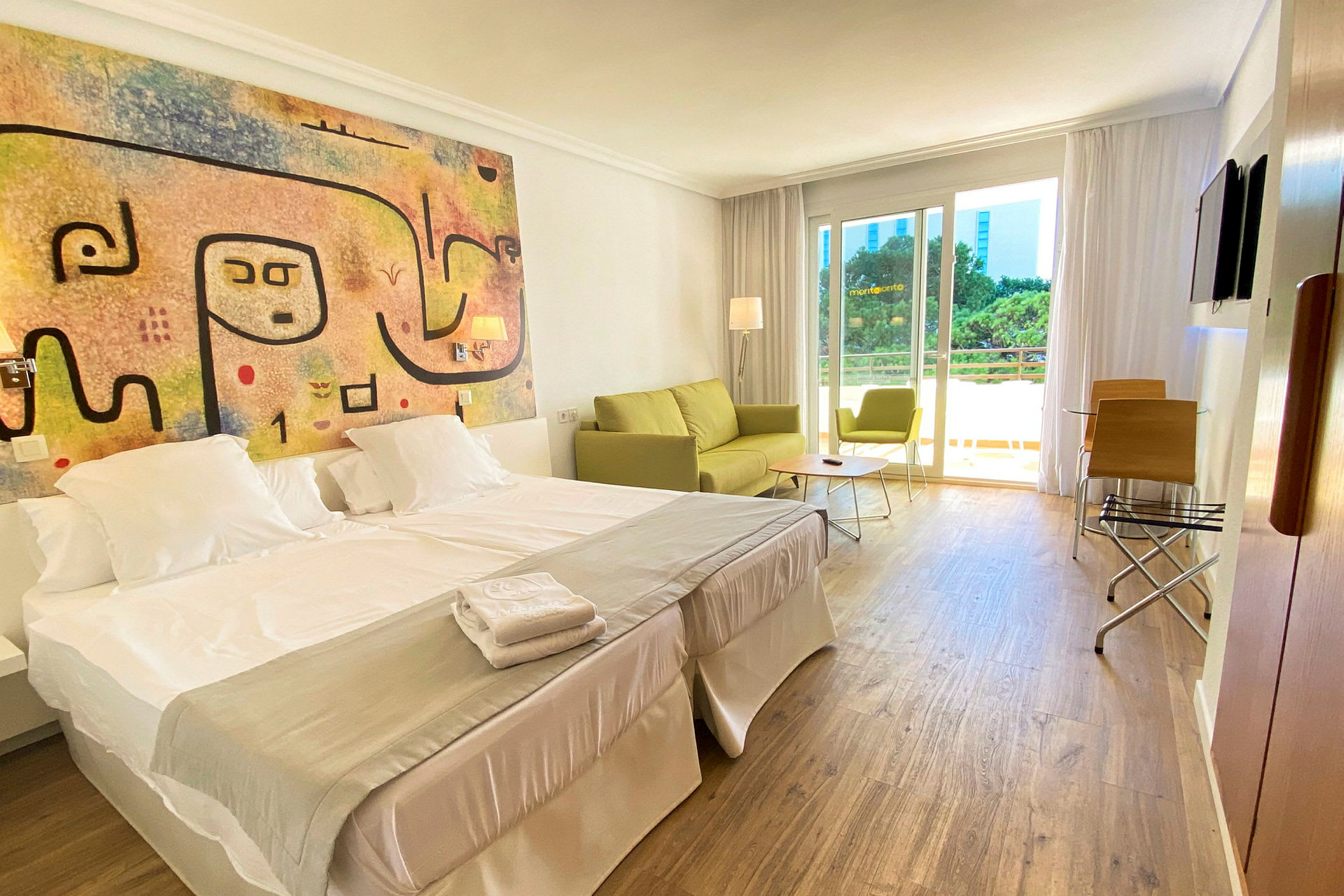 Et hotellrom med dobbeltseng, fargerikt veggkunst, og en sittegruppe med sofa og stol ved vinduet. (AI-generert tekst)