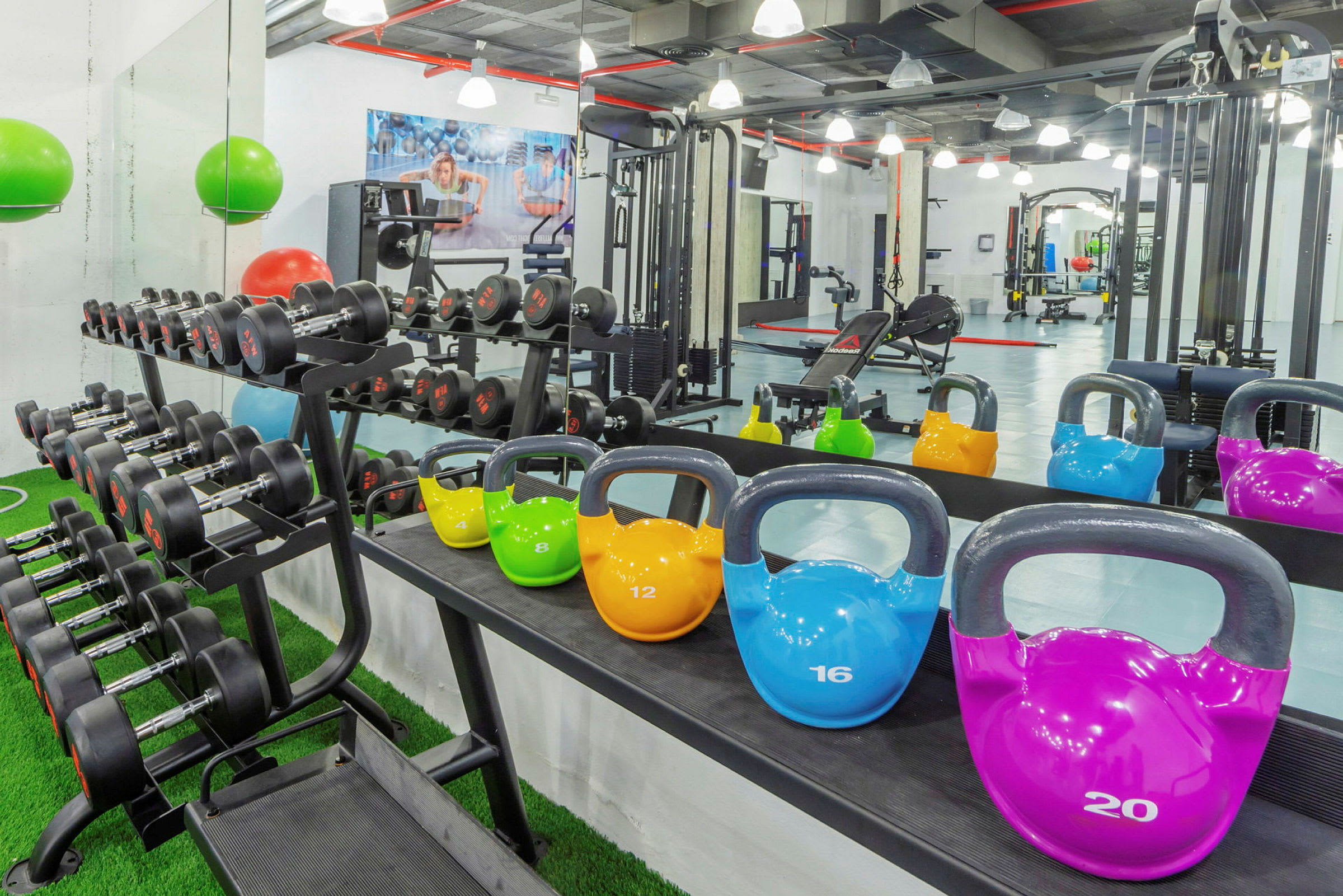 Bilde av et treningsstudio med manualer på et stativ og kettlebells i ulike farger plassert på en hylle, med treningsutstyr i bakgrunnen. (AI-generert tekst)