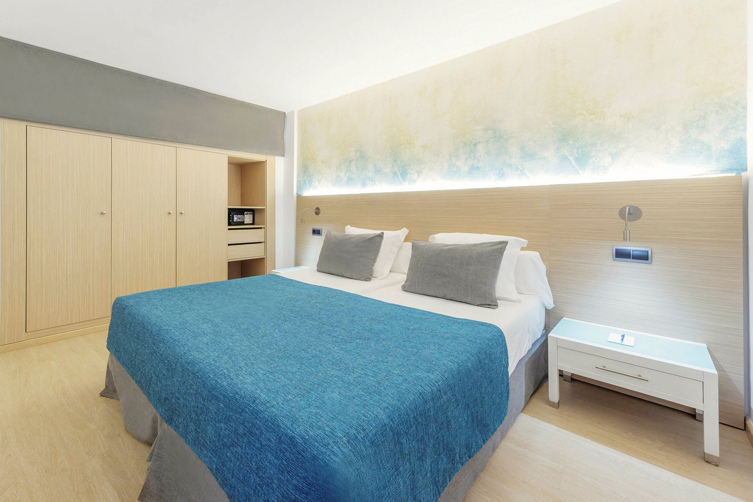 Hotellrom med dobbeltseng, blåt sengeteppe, hvite puter og minimalistisk innredning. (AI-generert tekst)