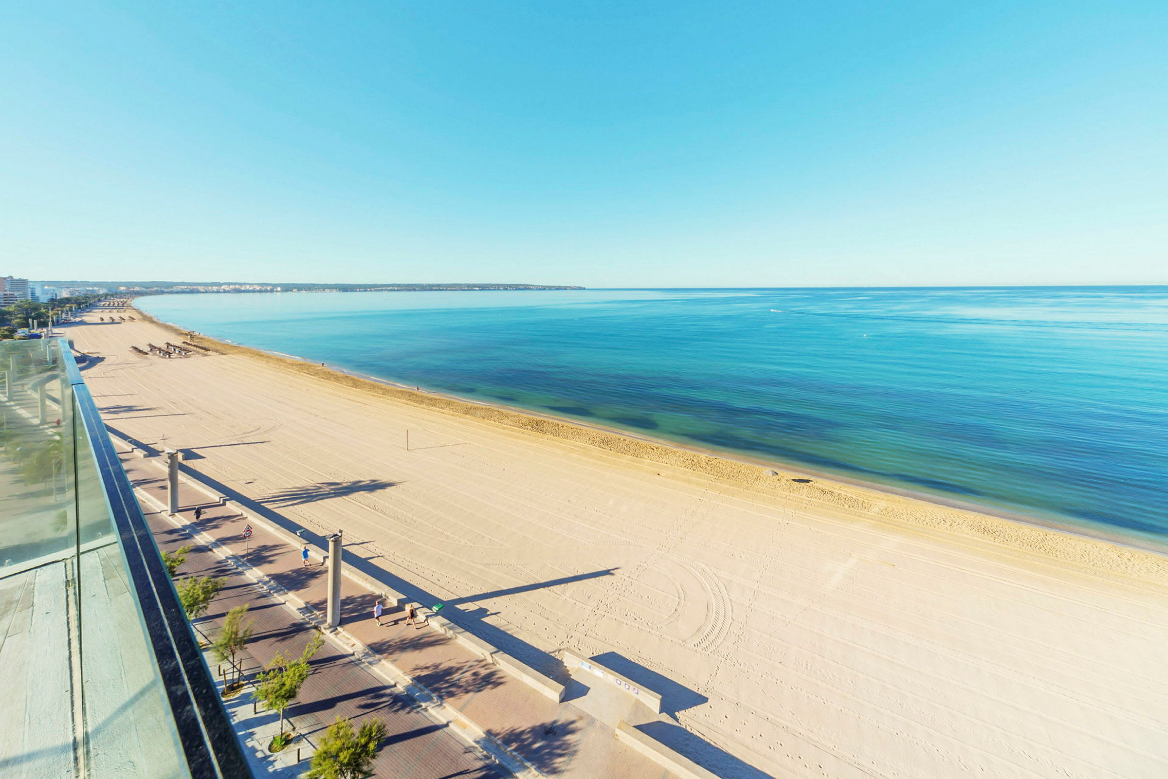 En lang sandstrand med klart blått hav og en promenade med gangvei og trær. (AI-generert tekst)