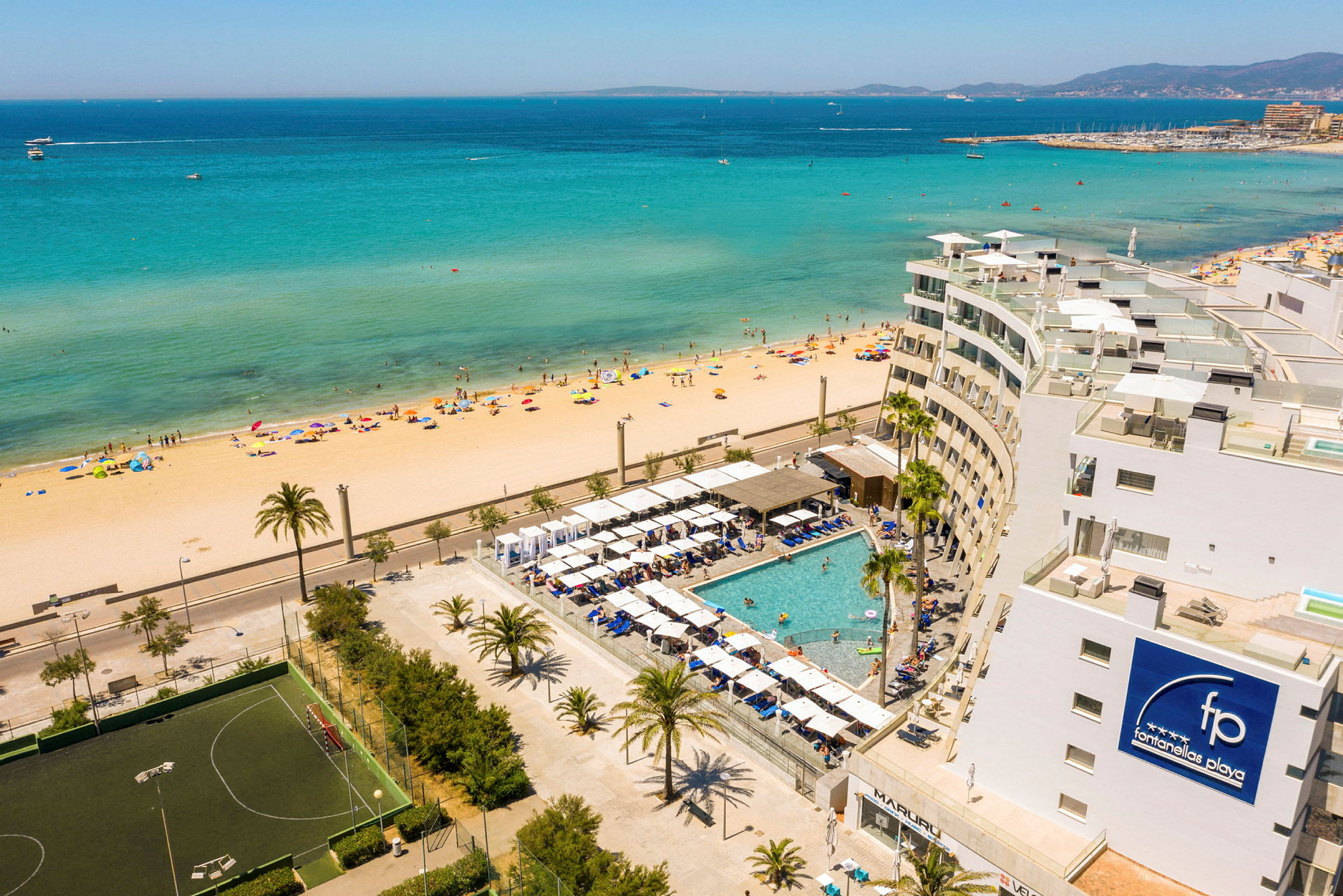 Utsikt over en strand med klart turkisblått hav og et hotellresort med basseng i forgrunnen. (AI-generert tekst)