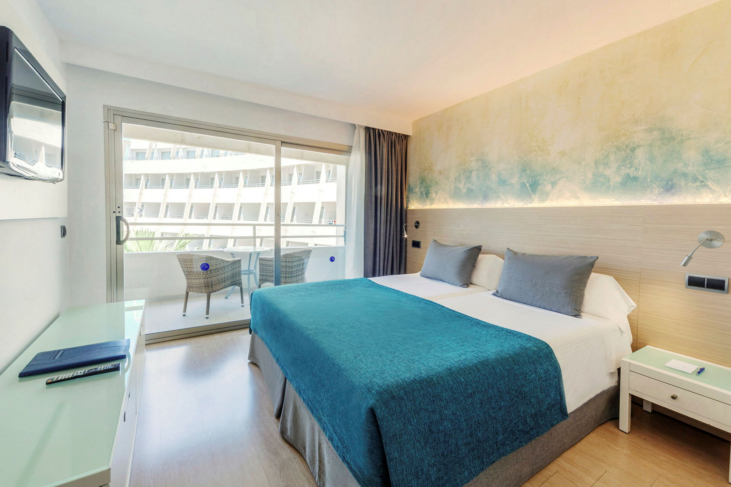 Et hotellrom med en dobbeltseng, blått sengeteppe og nattbord. En balkong med sittemøbler er synlig gjennom glassdøren. (AI-generert tekst)