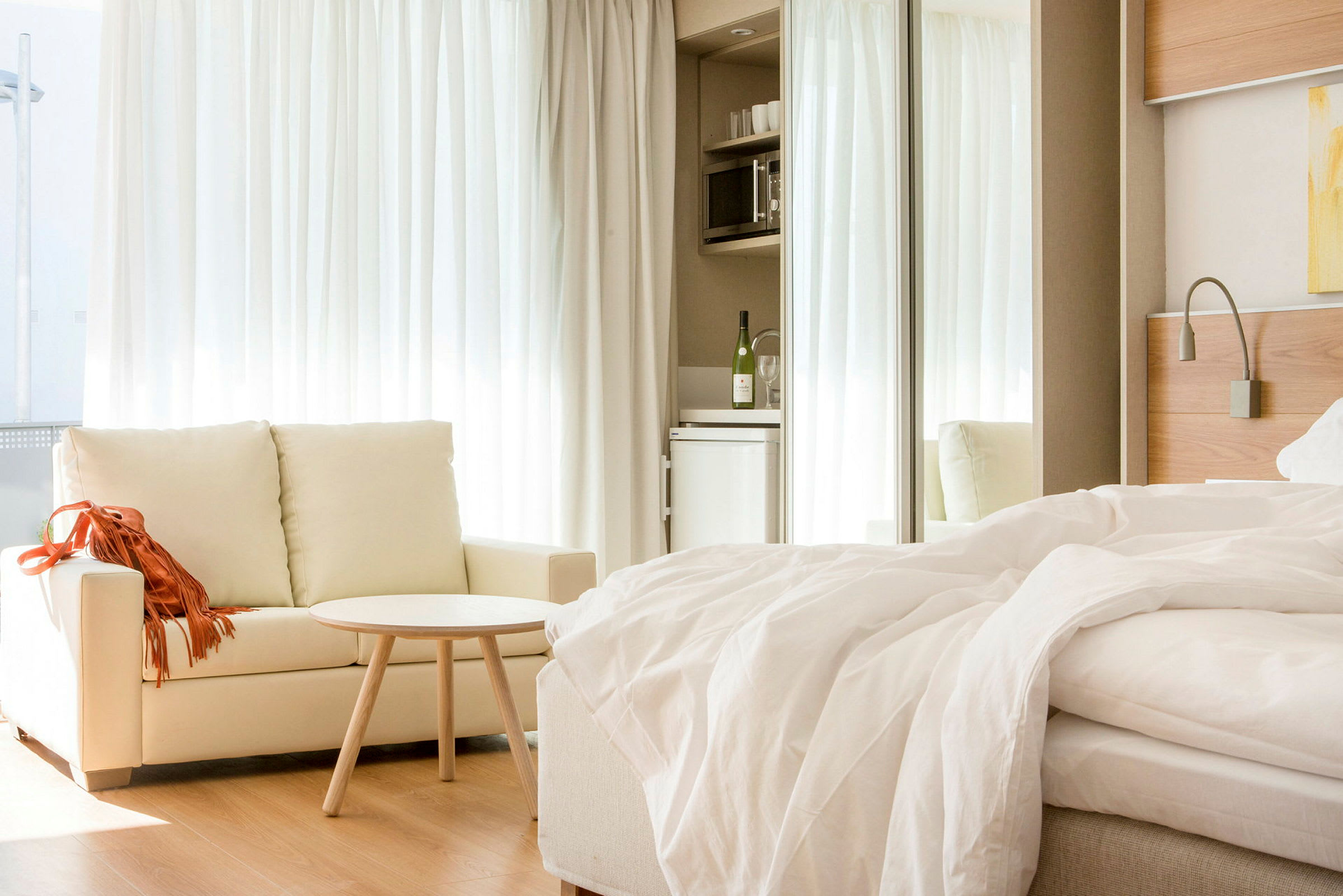 Et lyst hotellrom med en sofa, et lite bord og en oppredd seng. I bakgrunnen er det et minikjøkken med kjøleskap. (AI-generert tekst)