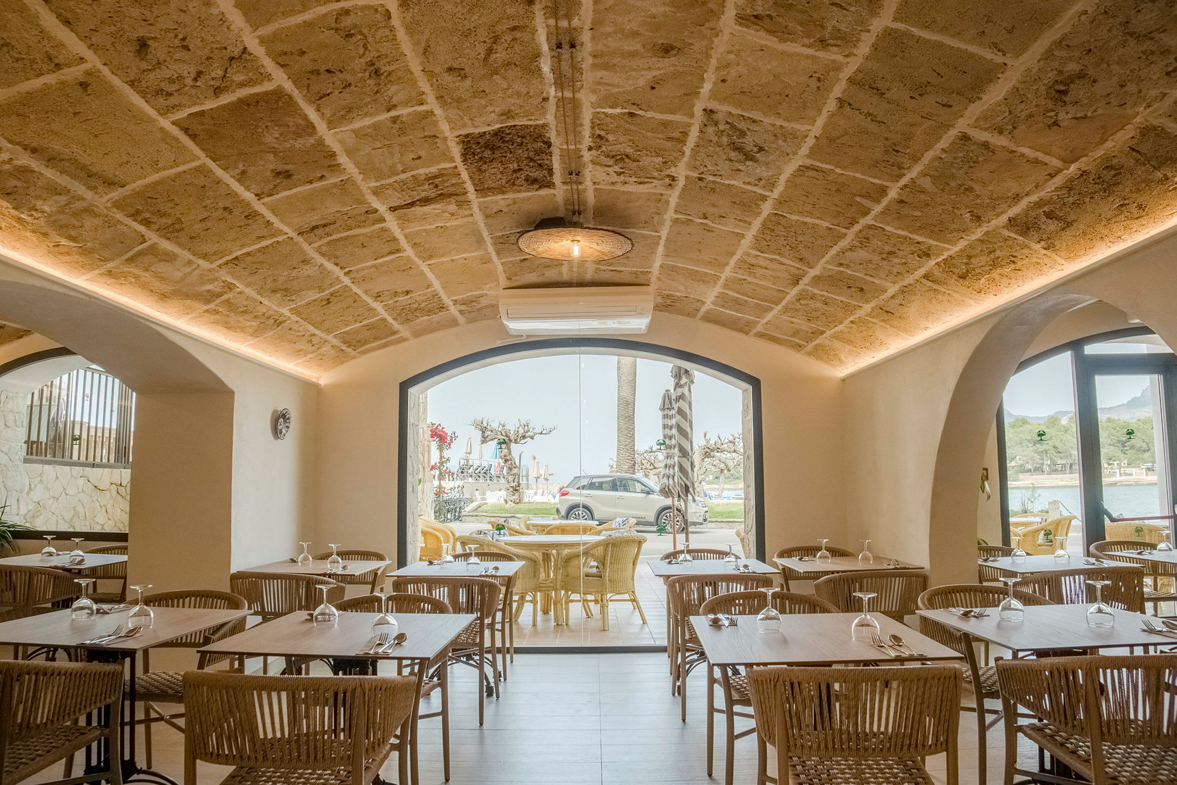 Interiør av en restaurant med steinvegger og hvelvet tak, bord og stoler i tre samt store vinduer som vender mot utsiktssteder utendørs. (AI-generert tekst)