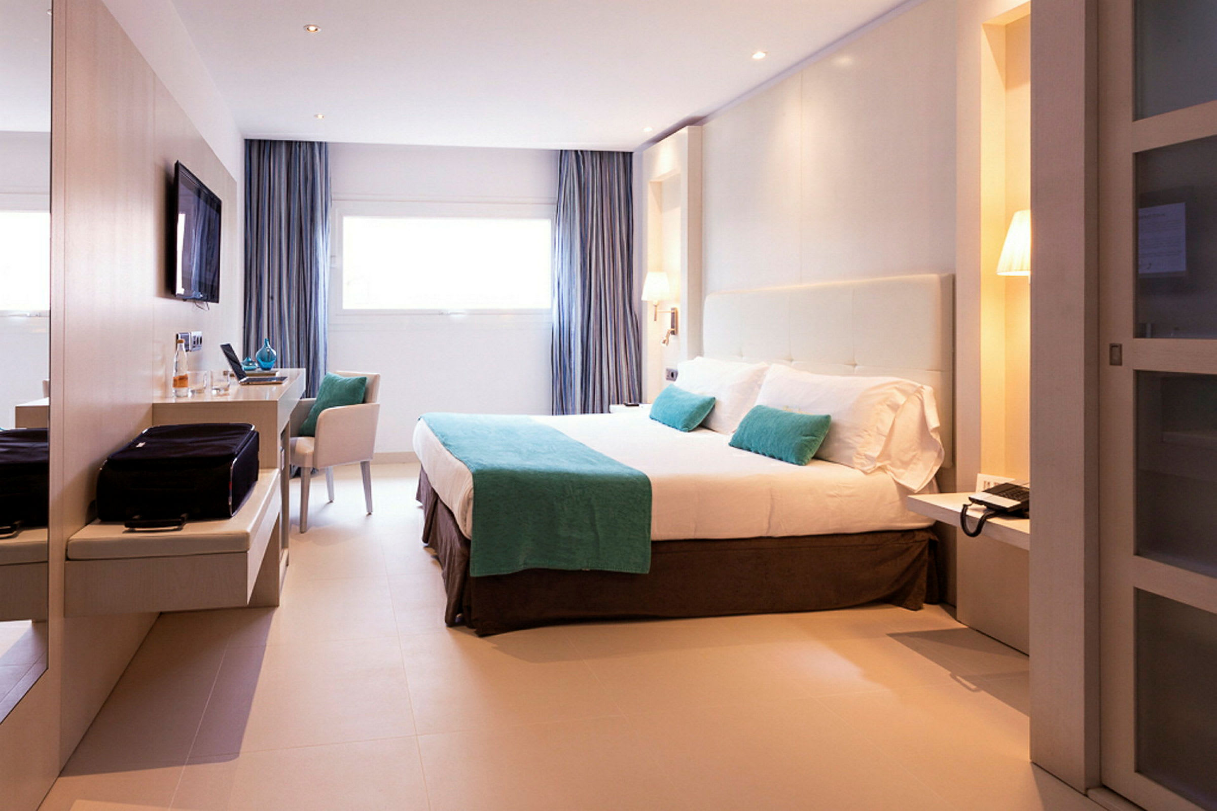Et moderne hotellrom med en stor seng, en TV på veggen og et skrivebord. Rommet er innredet i lyse farger med blå aksenter. (AI-generert tekst)