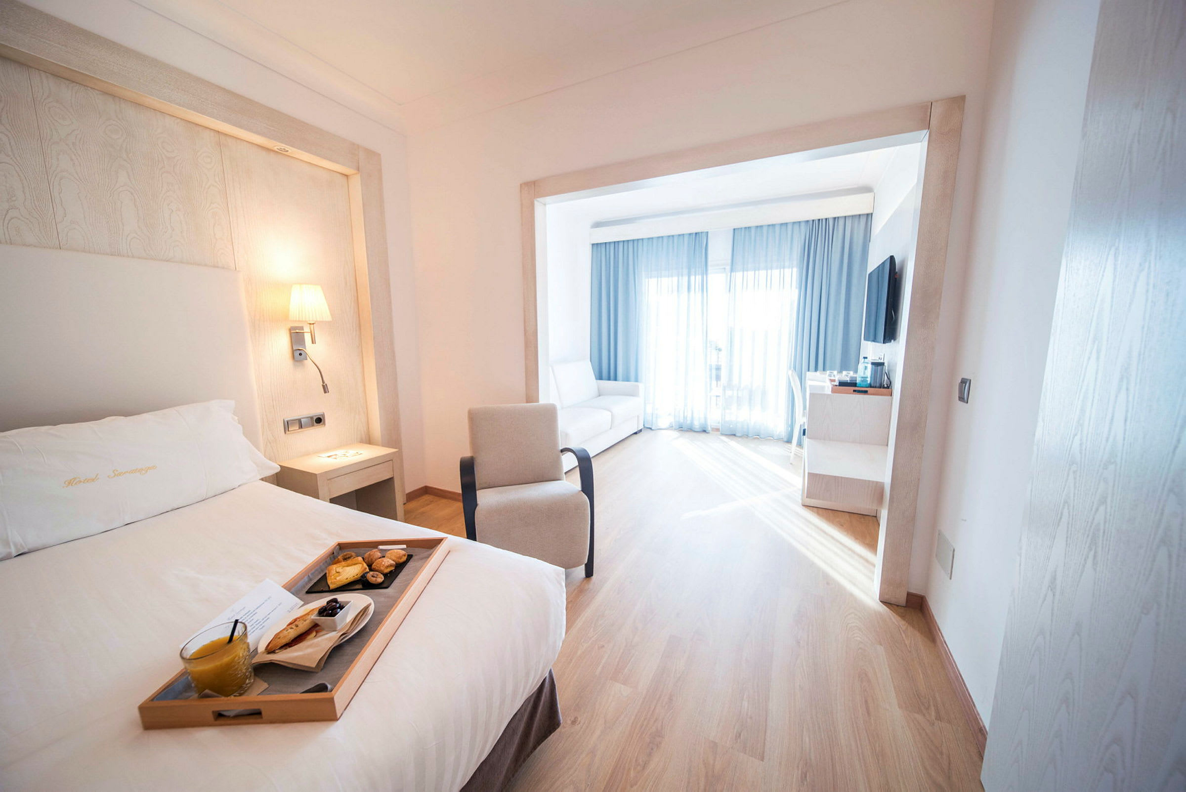 Et hotellrom med en seng og et brett som inneholder en frokost. Rommet har et stort vindu med lyse gardiner. (AI-generert tekst)