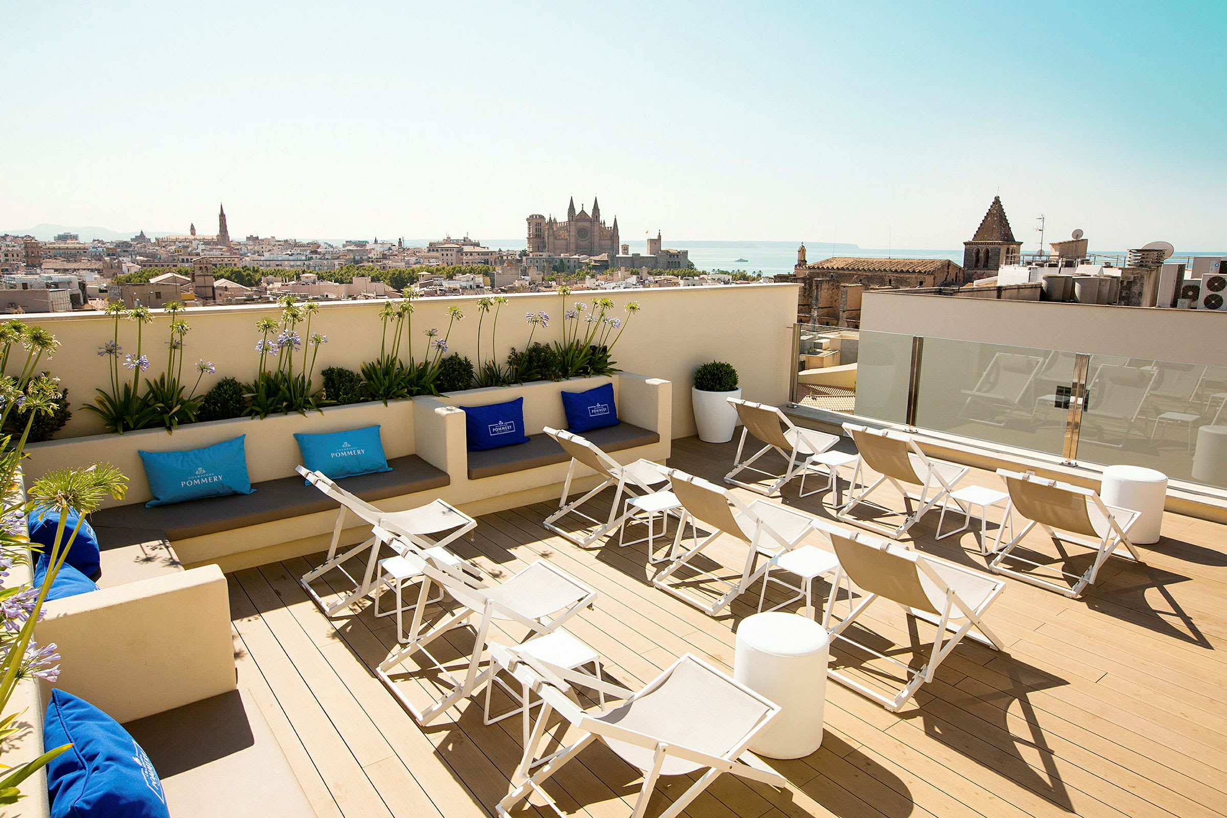 Utsikt fra en takterrasse med solsenger og sitteplasser, med en by og dens katedral i bakgrunnen. (AI-generert tekst)