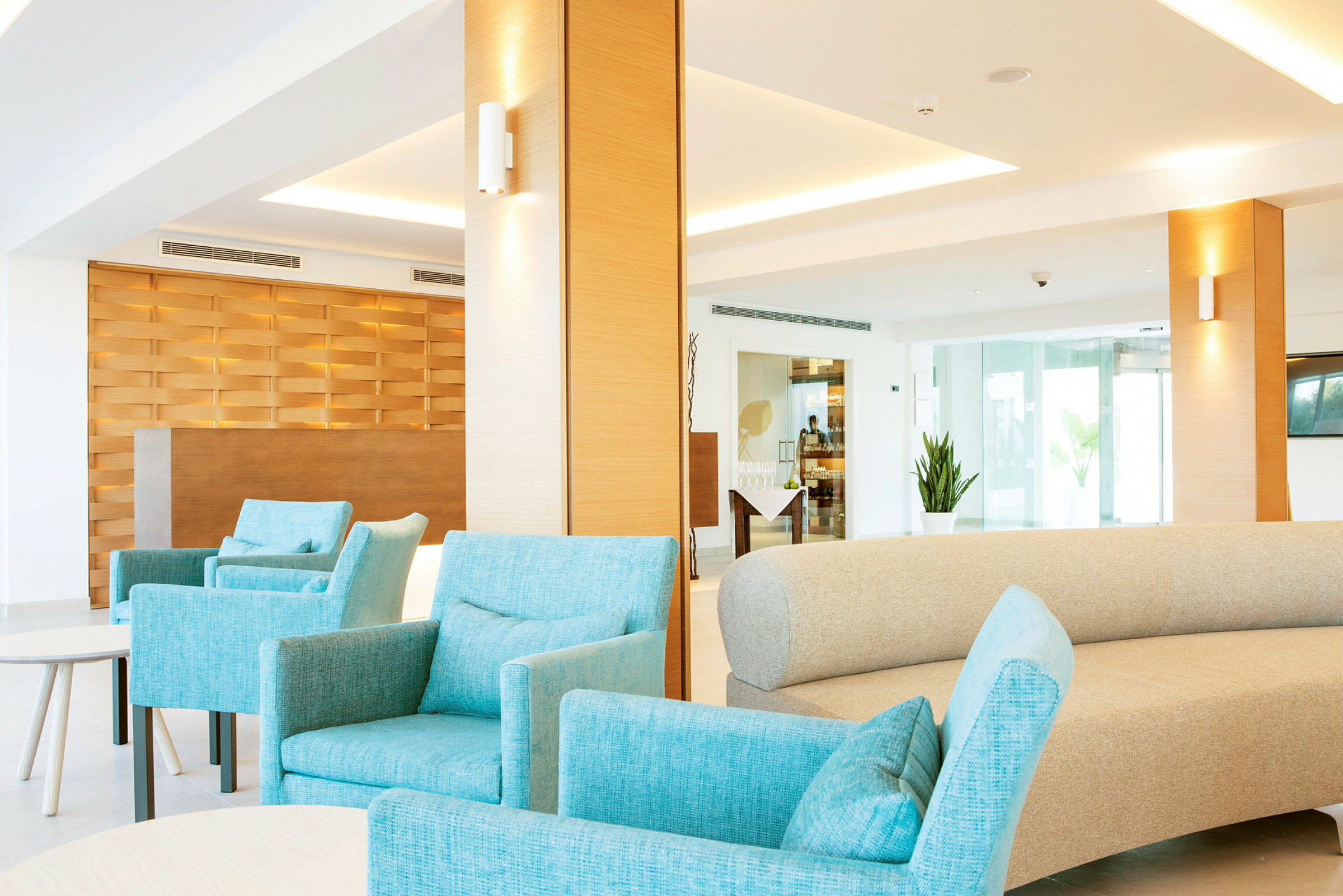 Interiør av en moderne lounge med turkise lenestoler og en beige sofa. Bakgrunnen viser en resepsjon med en trepanel og store vinduer. (AI-generert tekst)