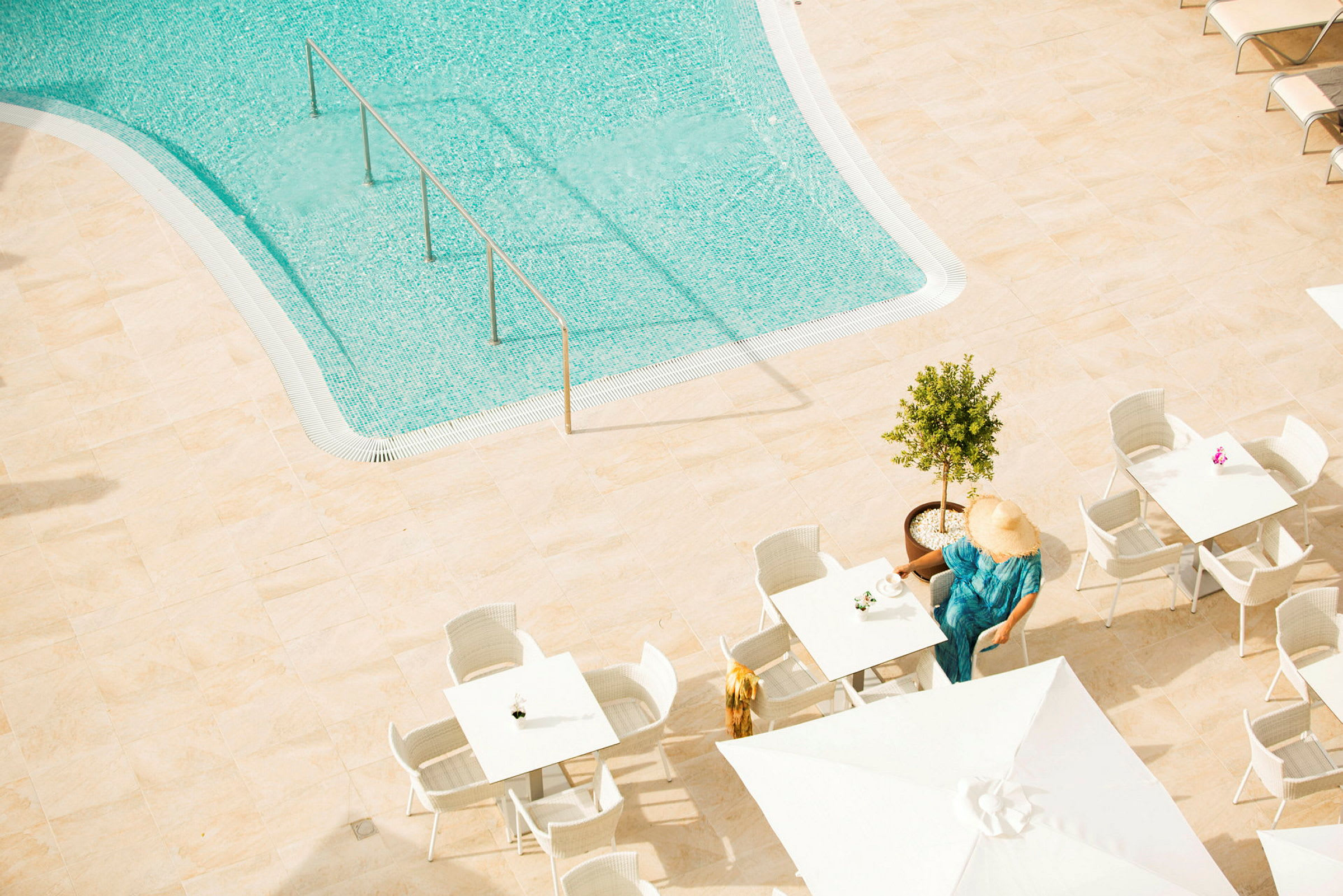 Utsikt over et basseng med klart vann. På en solterrasse er det bord og stoler, hvor en person sitter ved et bord med en kopp. Personens ansikt dekkes av en solhatt. (AI-generert tekst)