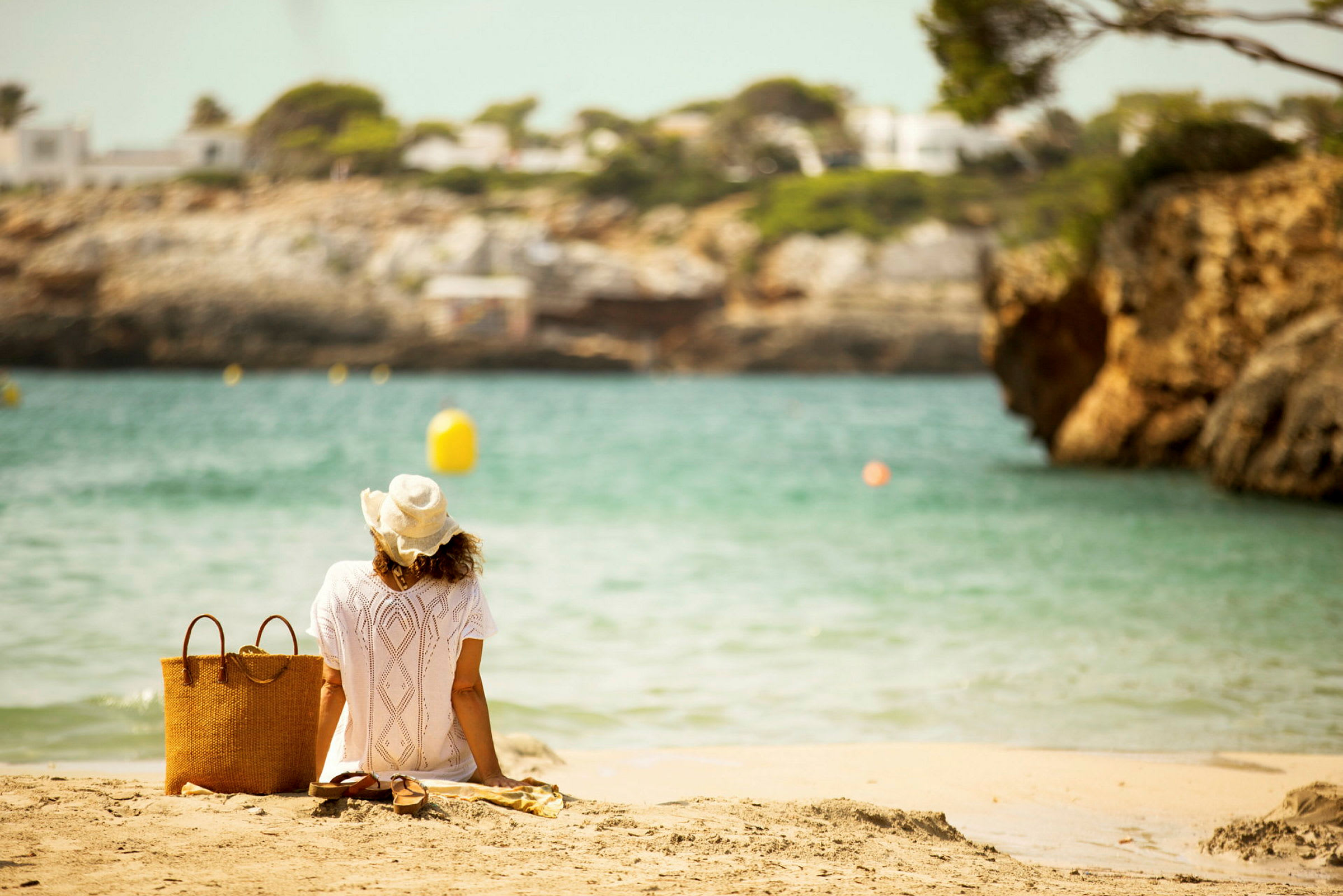 En person i en hvit kjole og hatt sitter på en strand og ser ut over vannet. Ved siden av personen er det en kurvveske og et par sandaler. (AI-generert tekst)