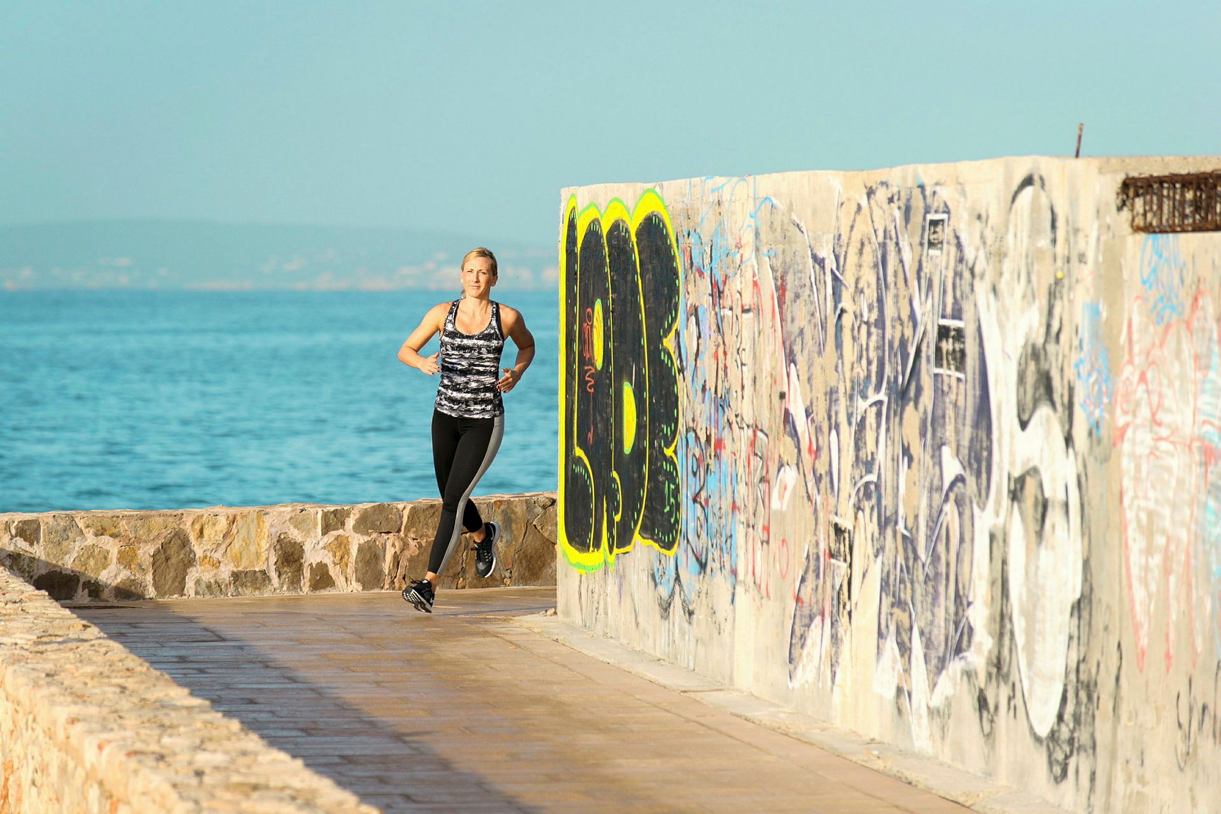 Person jogger langs en strandpromenade med havet i bakgrunnen og en vegg malt med graffiti. (AI-generert tekst)