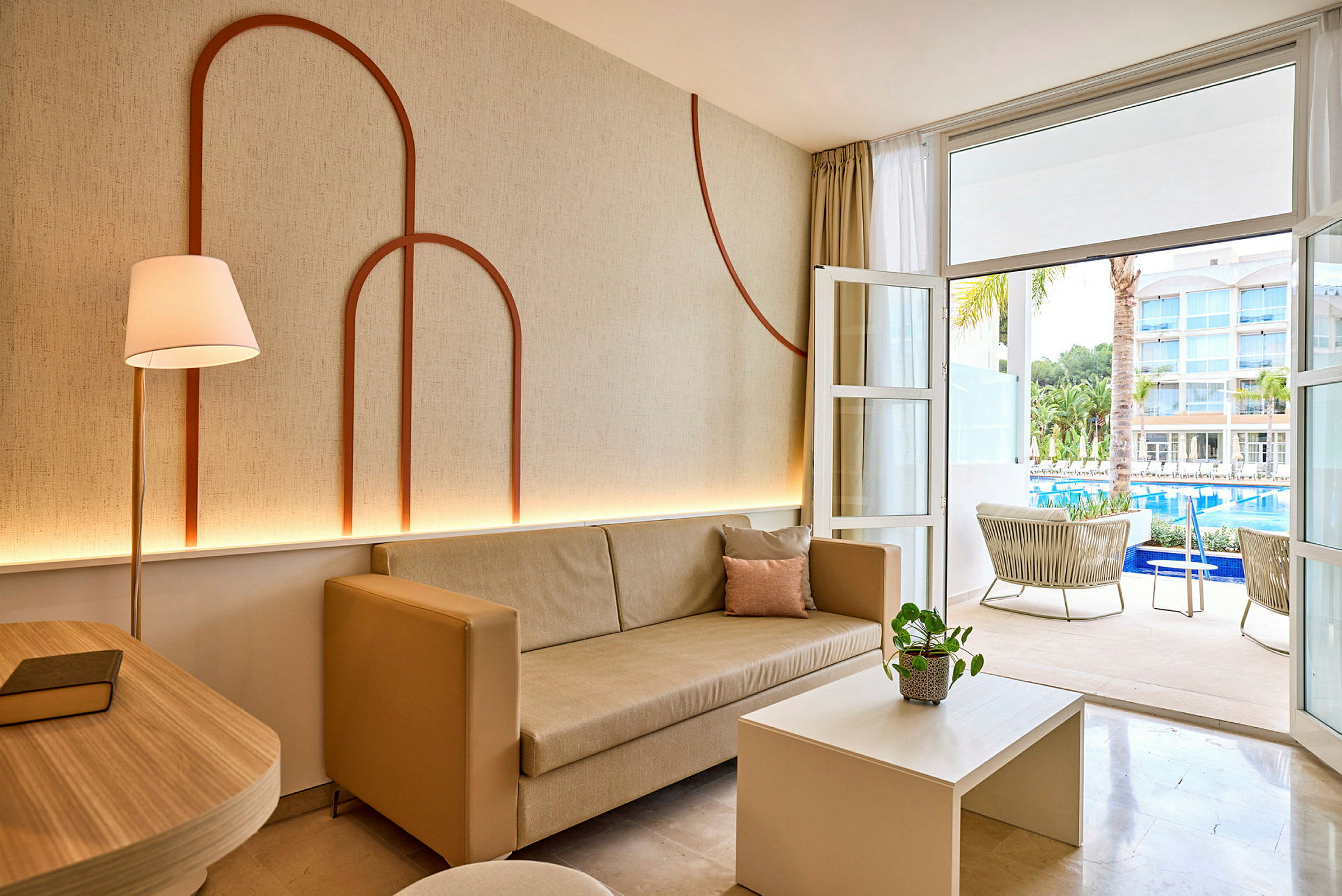 Stue med beige sofa og dekorert vegg, åpning mot terrasse med utsikt over basseng og hotellbygninger. (AI-generert tekst)