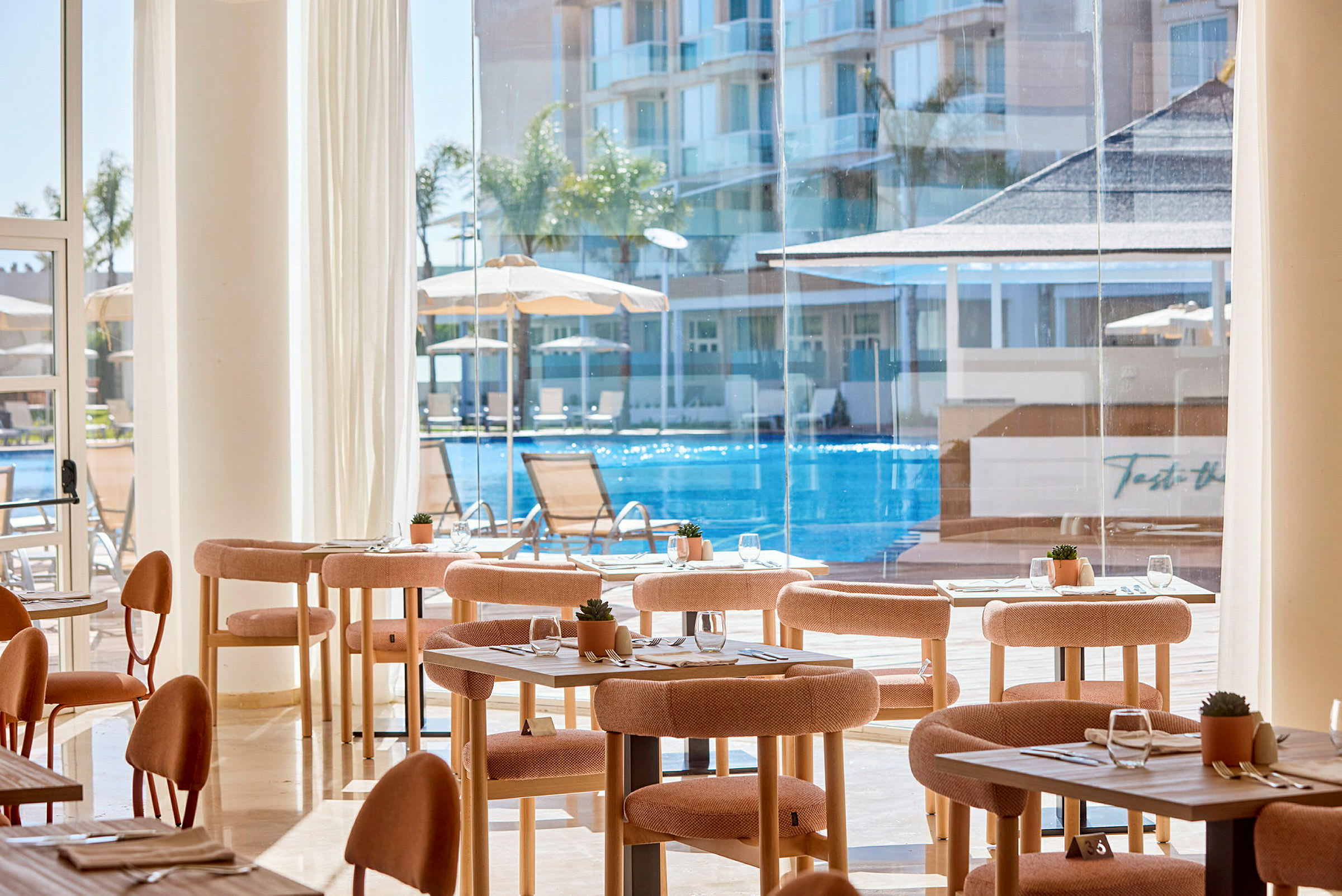 Interiør av en restaurant med bord og stoler, store vinduer med utsikt over et basseng og bygninger. (AI-generert tekst)