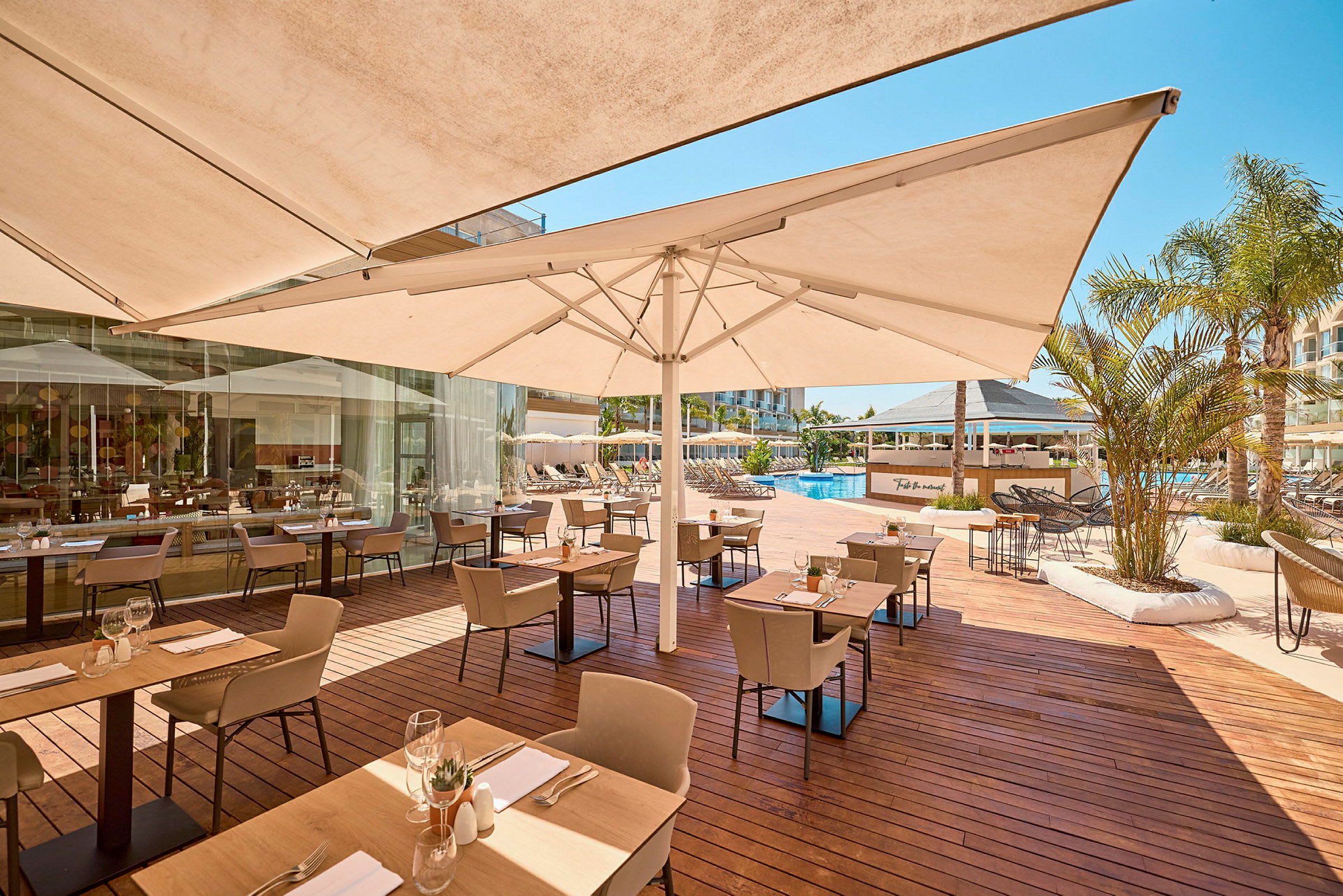 En utendørs restaurant ved et basseng, med bord og stoler på en treterrasse under parasoller. (AI-generert tekst)
