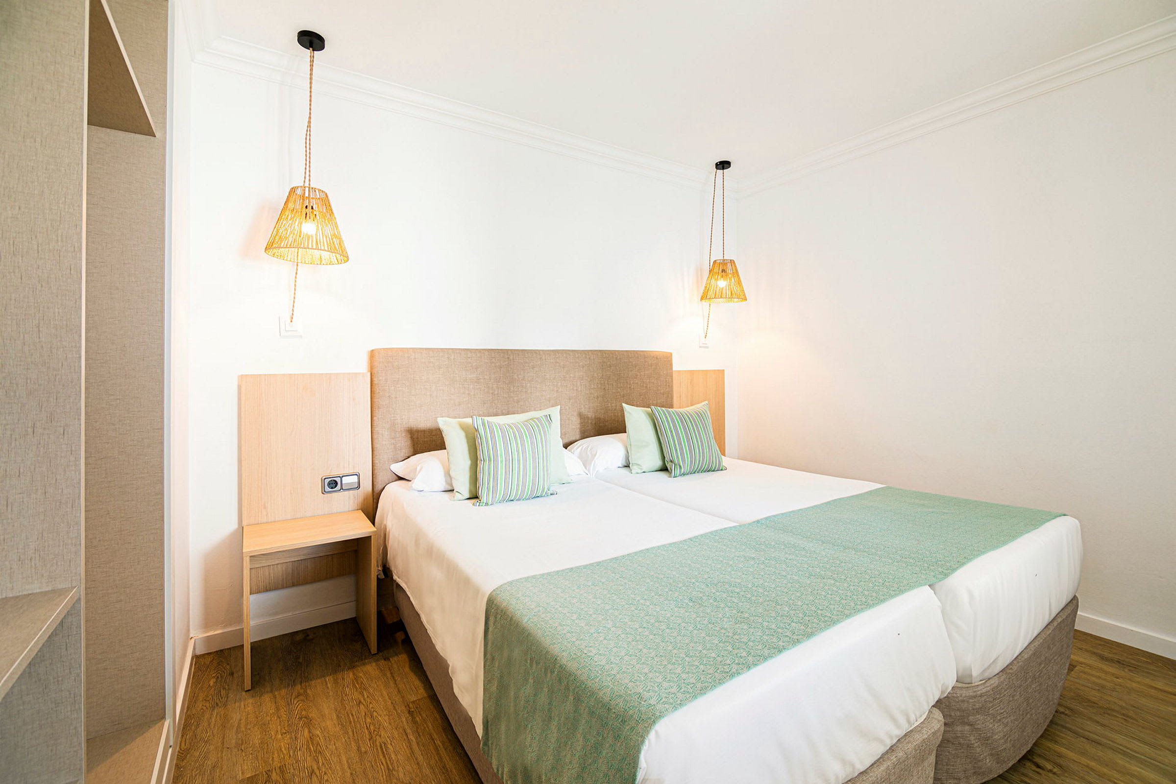 Et hotellrom med to oppredde enkeltsenger, grønne sengetepper og puter samt to hengende lamper. (AI-generert tekst)
