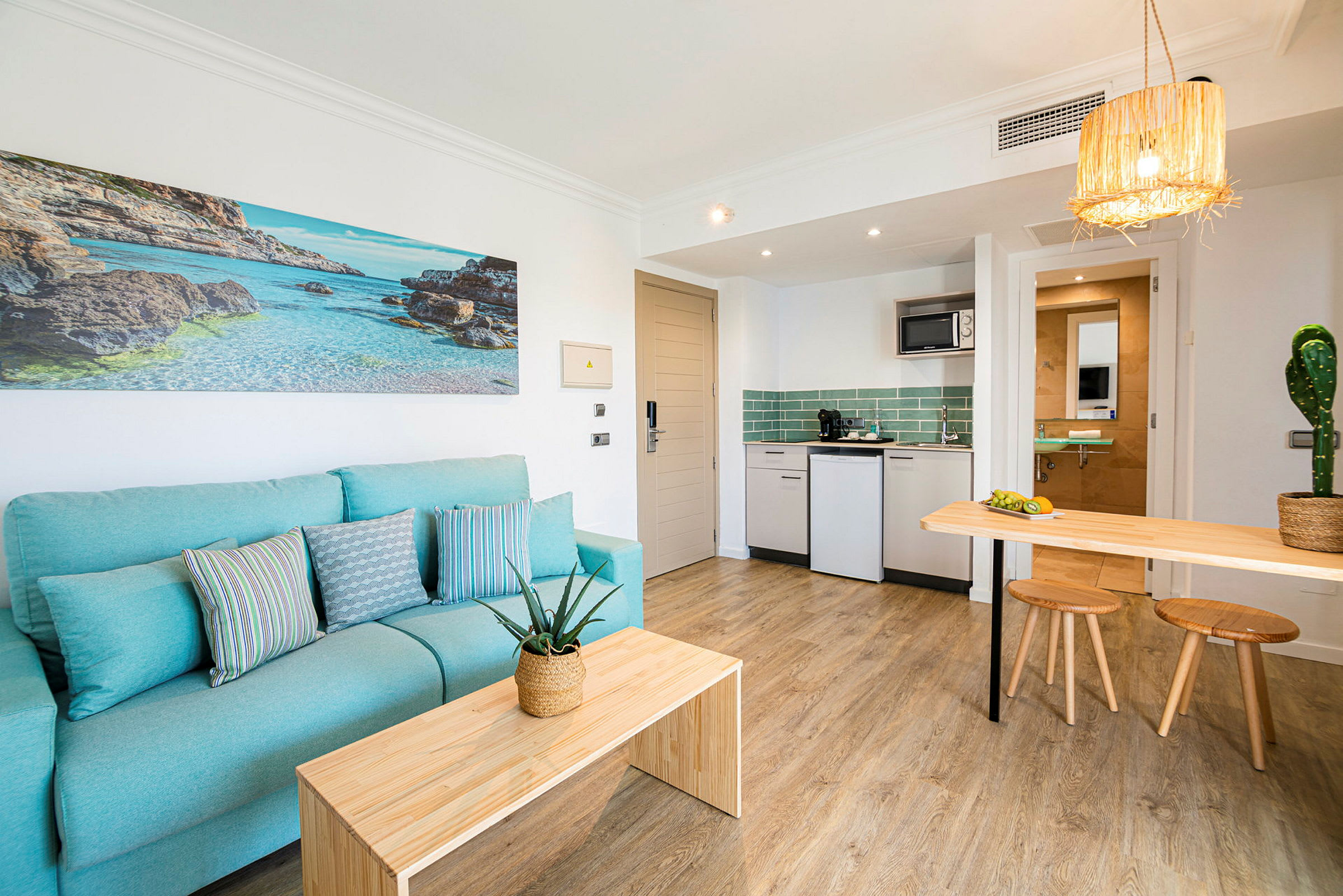 Et hotellrom med lyseblå sofa og en moderne kjøkkenkrok. Veggdekorasjon som viser en strand. (AI-generert tekst)