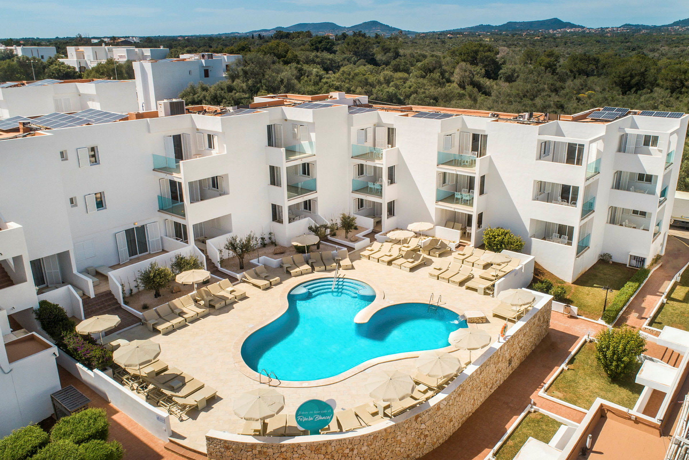 Et hotellområde med et utendørsbasseng og solsenger omgitt av hvit arkitektur. (AI-generert tekst)