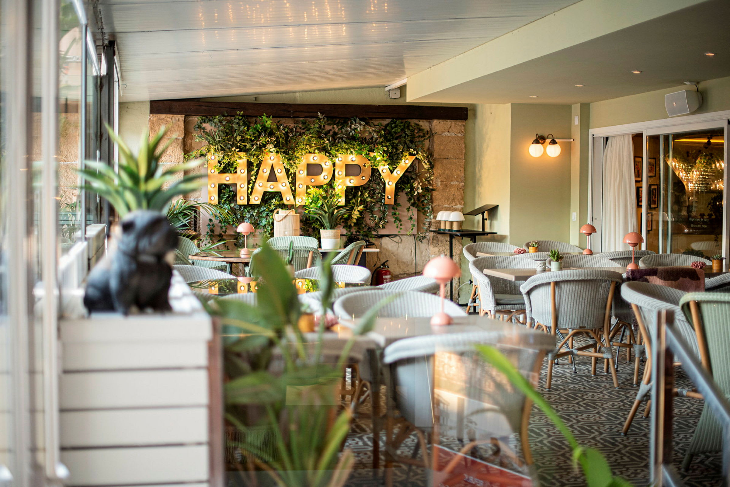 Interiør av en restaurant eller kafe med grønne planter, belysning og teksten 'HAPPY' på veggen. (AI-generert tekst)