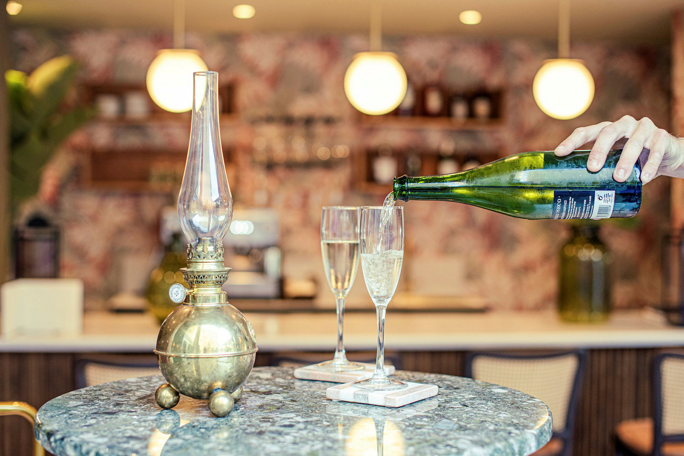 En antikk oljelampe på et bord ved siden av to champagneglass. En hånd heller drikke i ett av glassene. (AI-generert tekst)