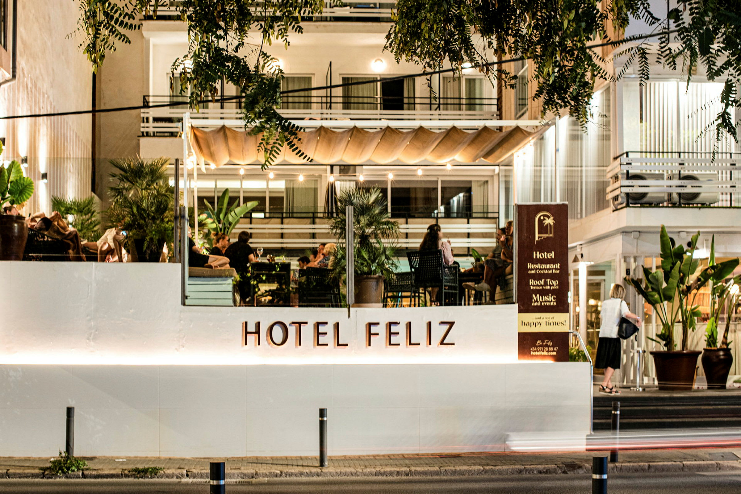 Hotel Feliz med opplyst fasade og terrasse der folk sitter. (AI-generert tekst)
