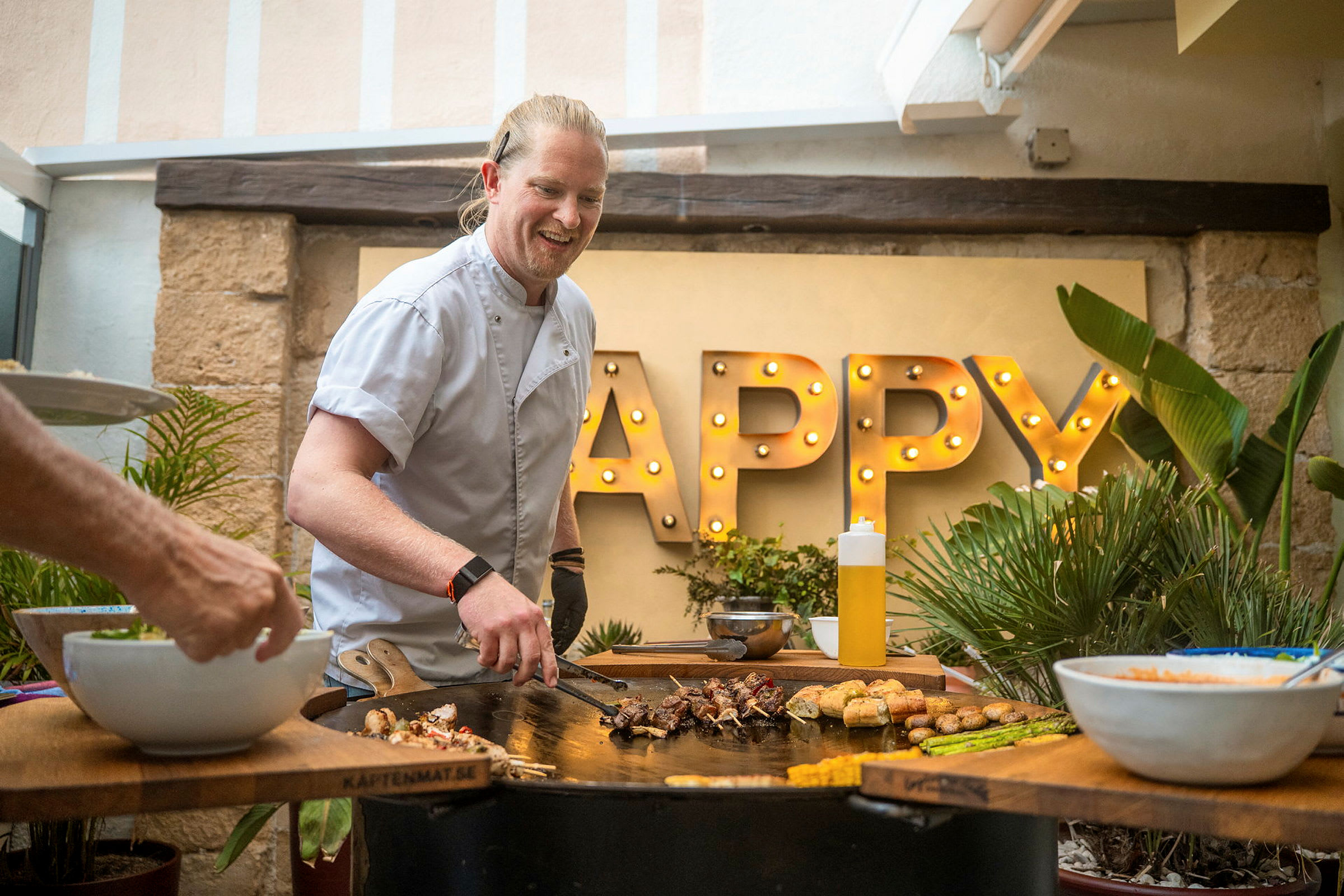 Person tilbereder mat på en grill med ordet 'Happy' opplyst i bakgrunnen. (AI-generert tekst)