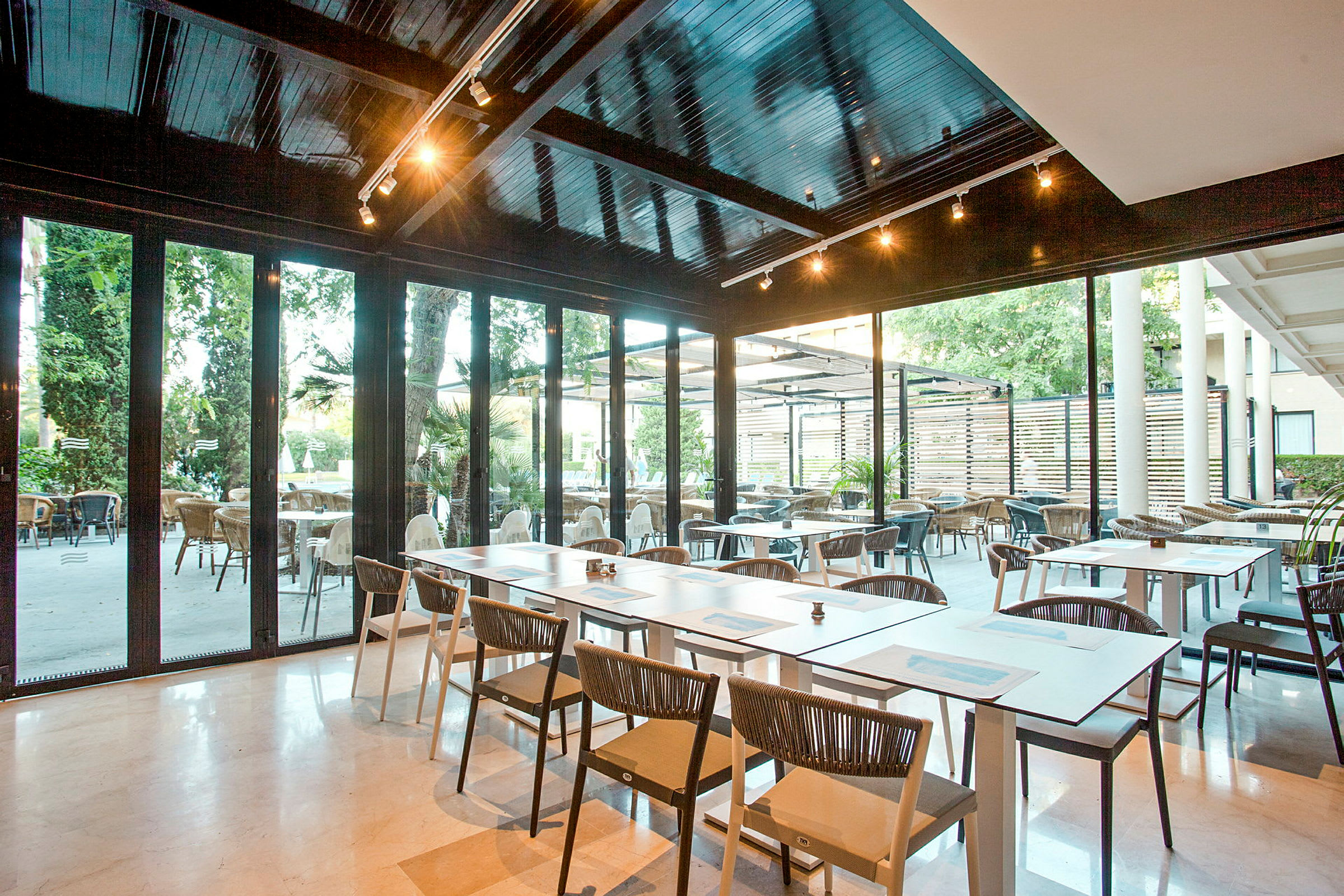 En moderne restaurant med sitteplasser både innendørs og utendørs. Bord og stoler er plassert på et lyst gulv, med glassvegger som gir utsikt over uteområdet. (AI-generert tekst)