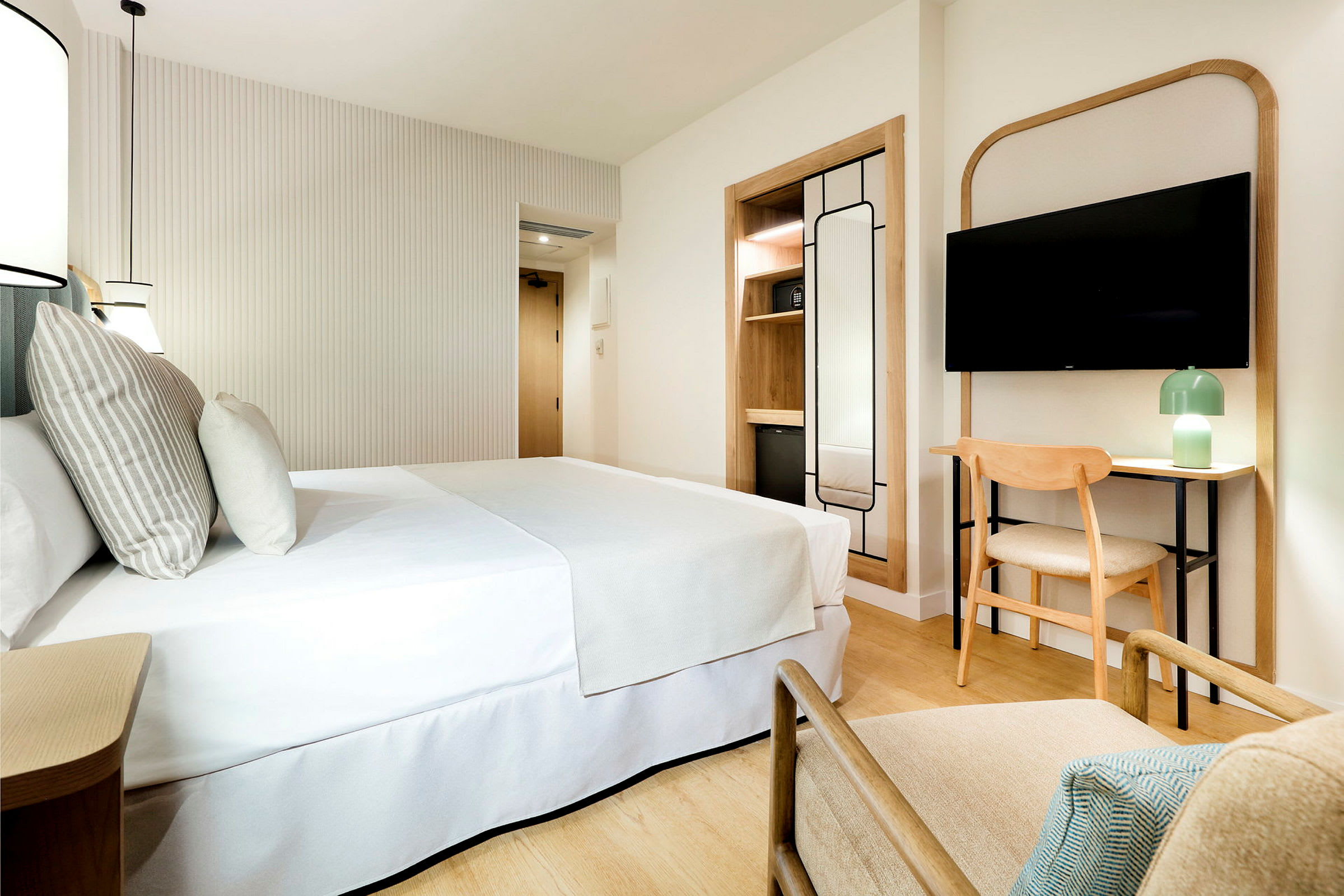 Et moderne og lyst hotellrom med en dobbeltseng, en TV på veggen, en lenestol, et skrivebord med stol, og en minimalistisk innredning. (AI-generert tekst)