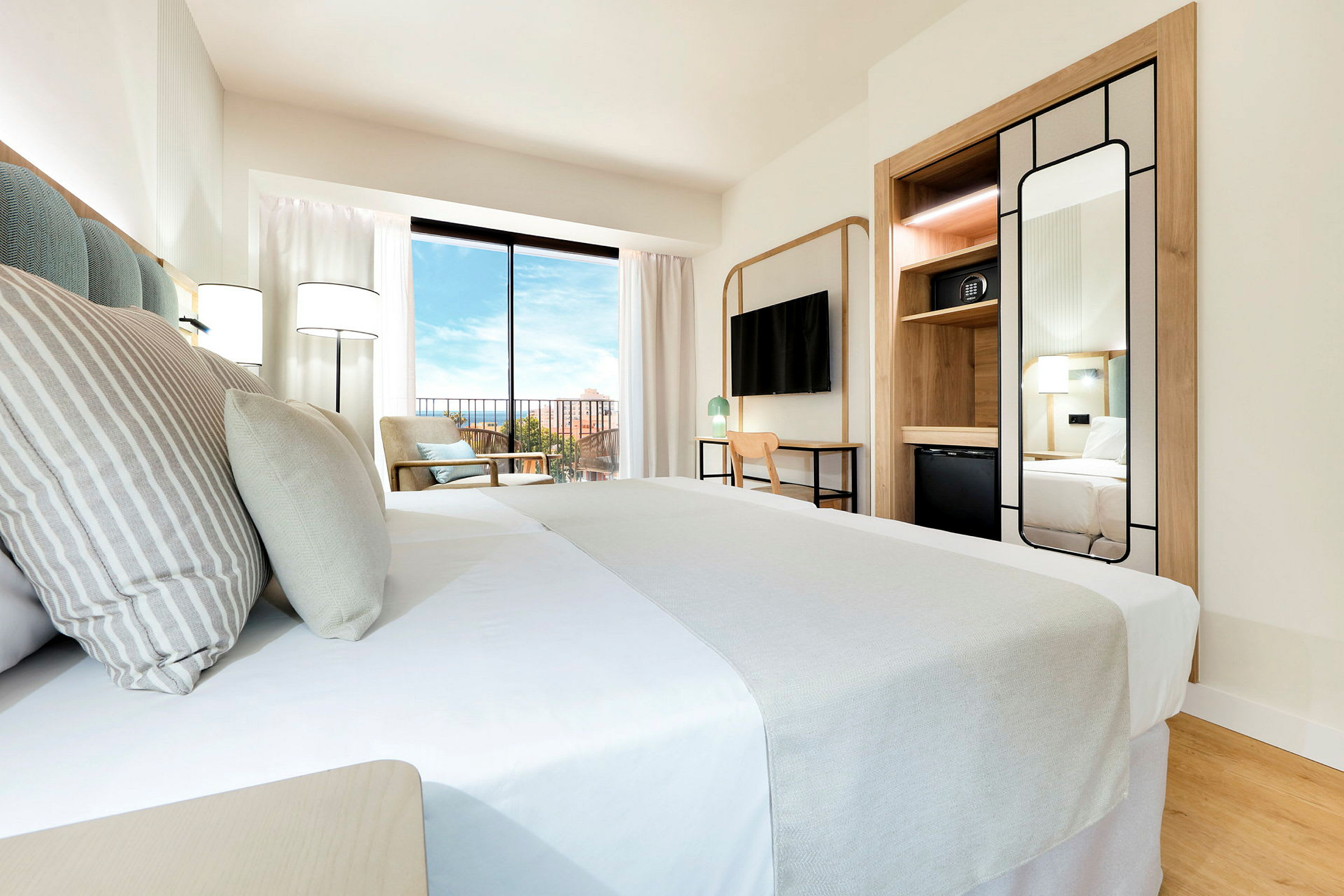 Et hotellrom med en oppredd seng i forgrunnen, et speil og oppbevaring på siden, samt en stor skyvedør som vender ut mot en balkong. (AI-generert tekst)