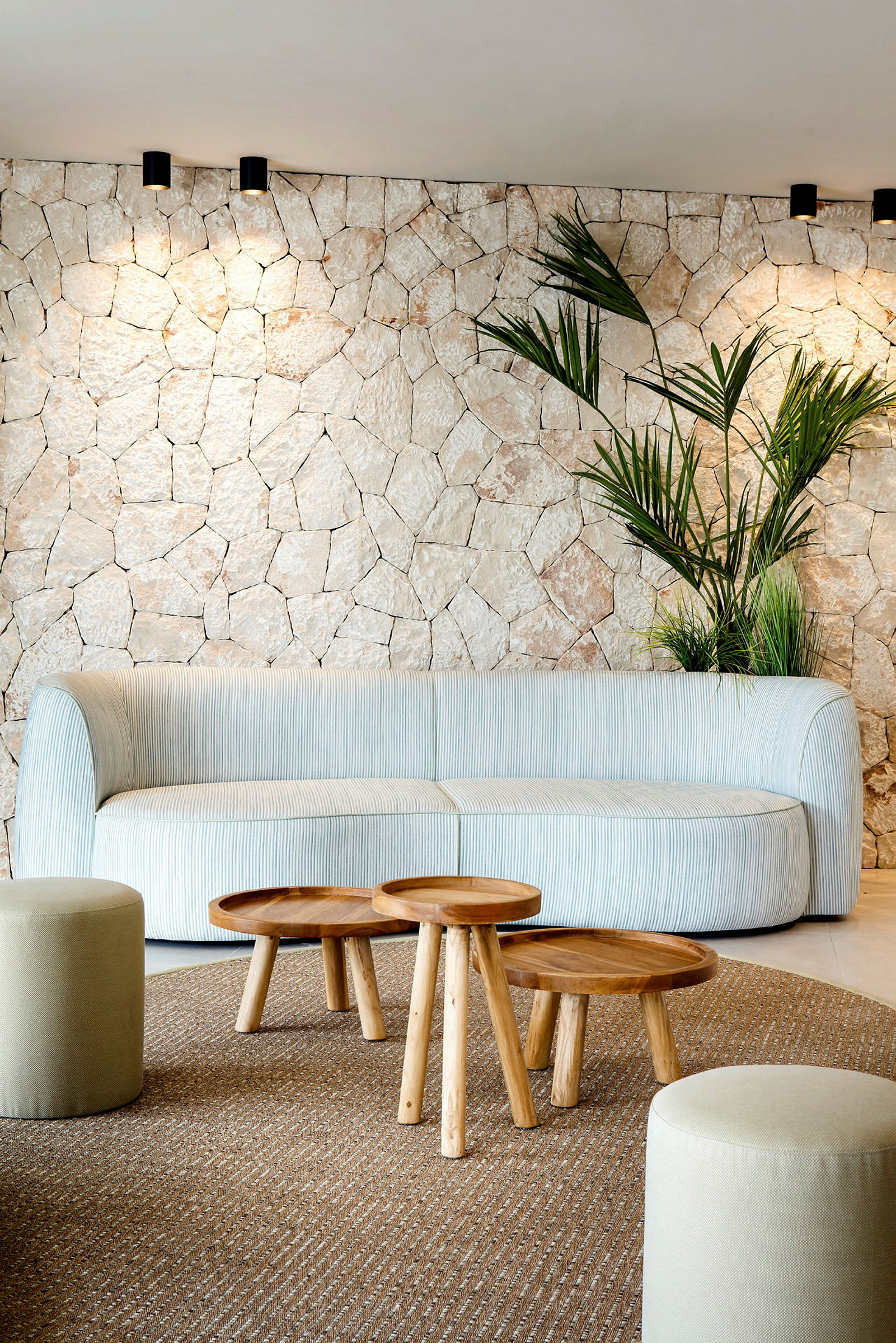 Et lyst stueinteriør med en beige sofa, små trebord og dekorative planter foran en vegg med steinstruktur. (AI-generert tekst)
