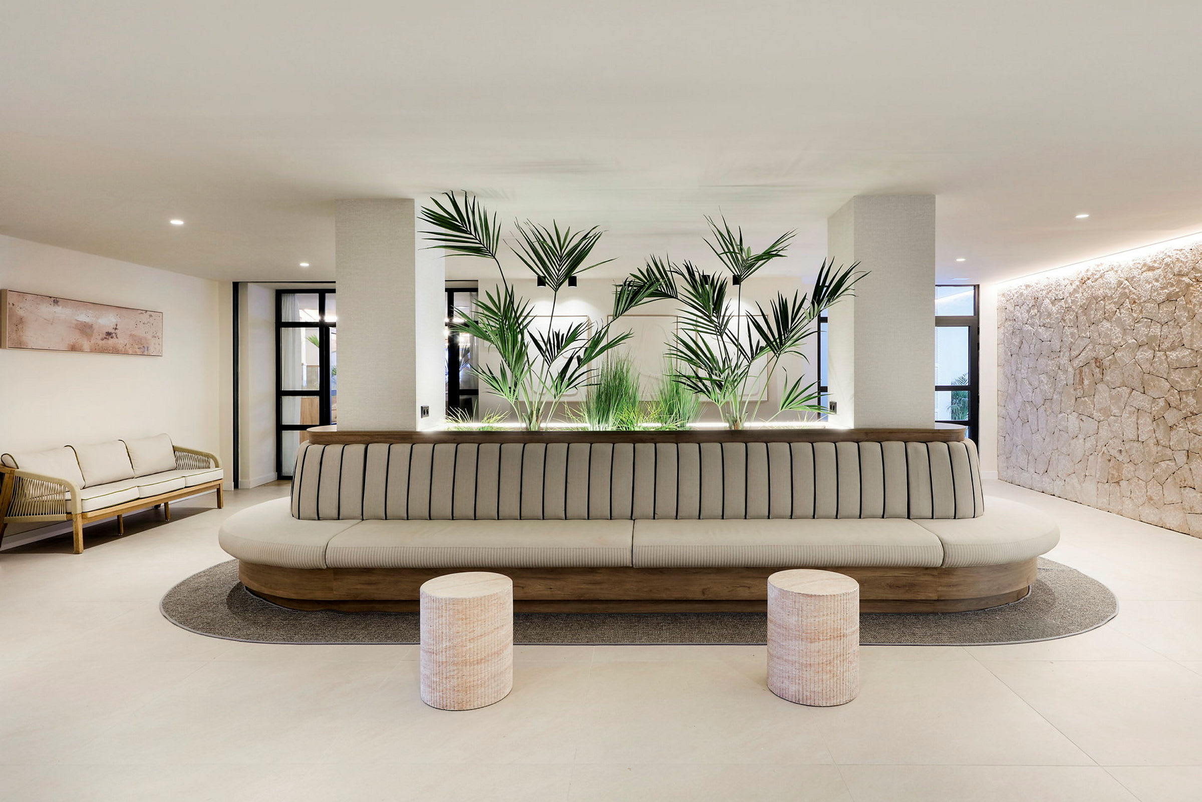 Et moderne venterom eller lounge med en lang, polstret sofa, to runde krakker foran, og grønne planter som dekorasjon i bakgrunnen. (AI-generert tekst)