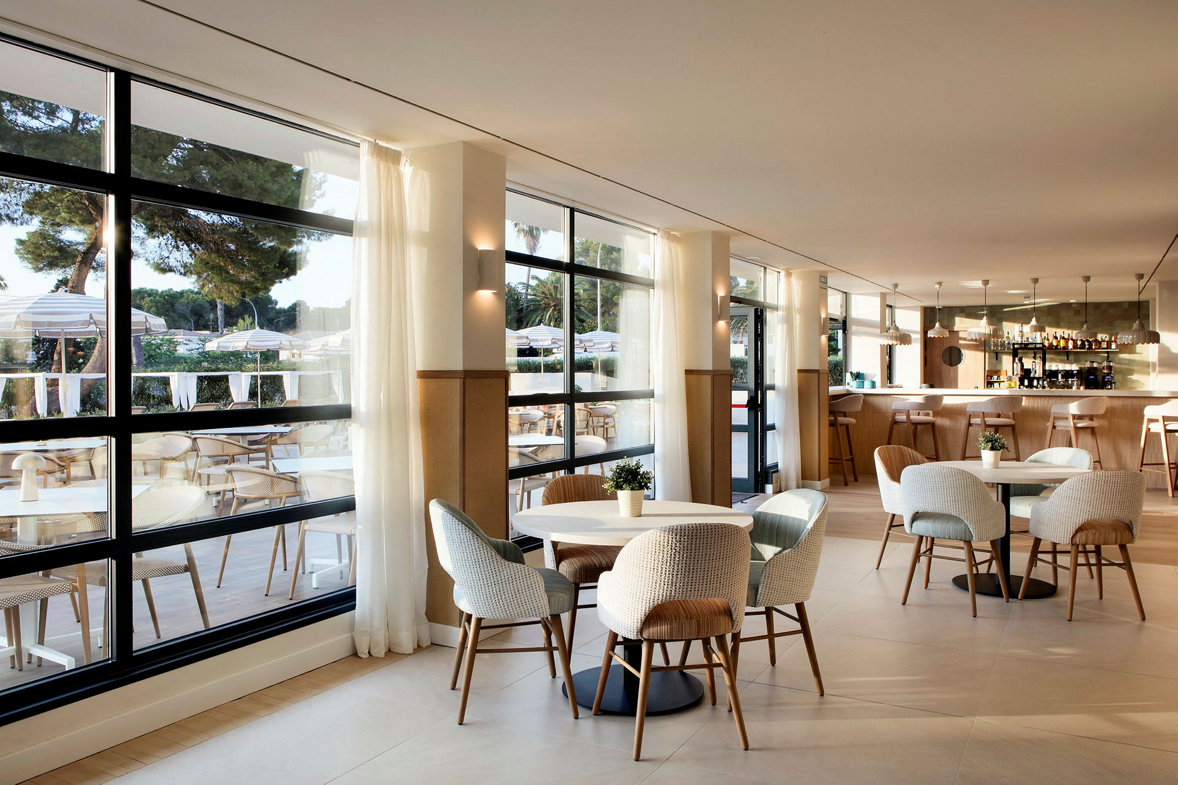 En moderne restaurant med store vinduer, lyse møbler og en innbydende innredning. Utsikt mot en terrasse med bord og parasoller. (AI-generert tekst)