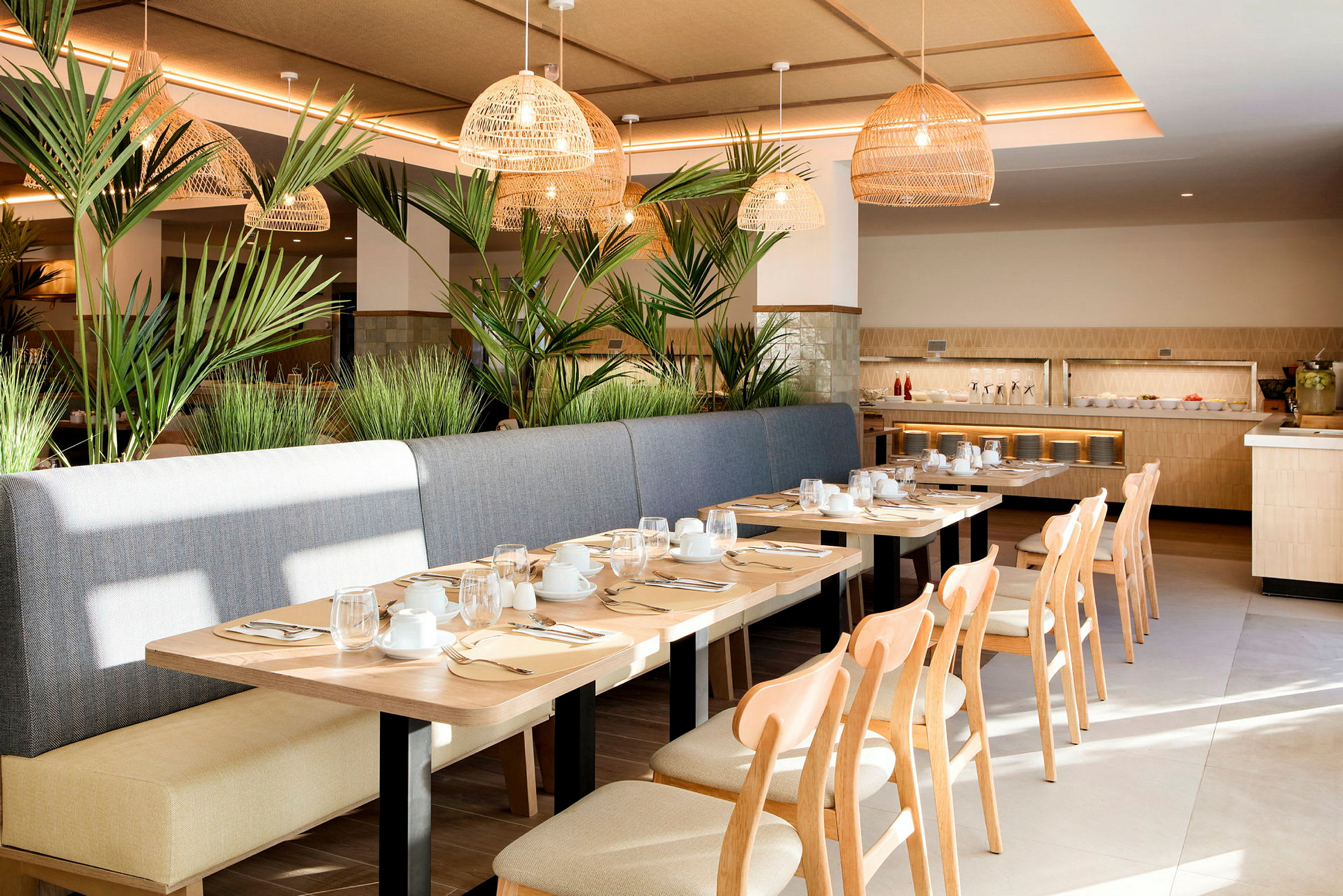 En lys og moderne restaurant med trebord, stoler og benk av stoff. Dekorert med grønne planter og flettede taklamper. Porselen og glass er dekket på bordene. (AI-generert tekst)