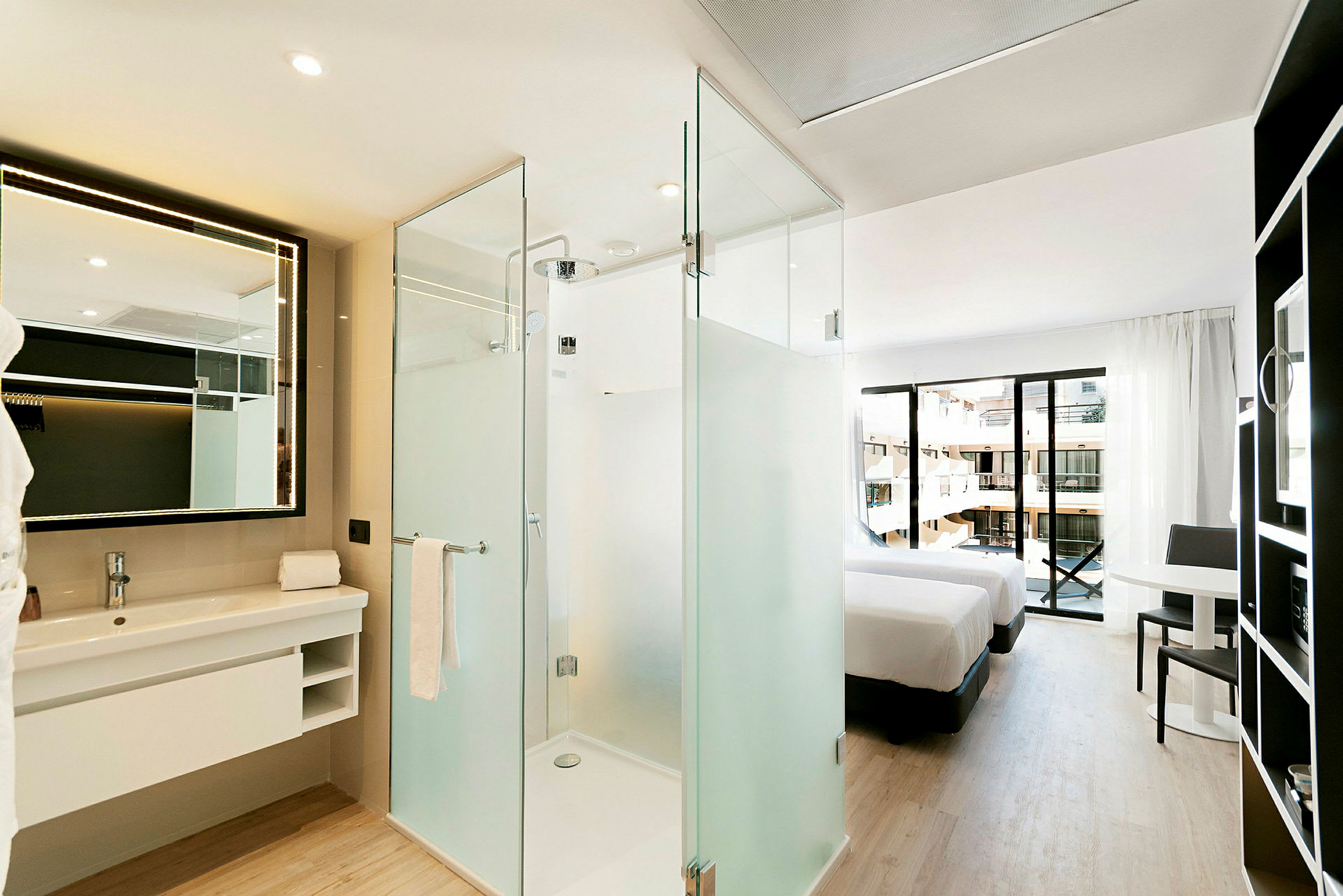 Et moderne hotellrom med soverom og bad. Badet har en dusj med glassvegger og et stort speil med belysning. Soverommet har to senger og tilgang til en balkong med utsikt. (AI-generert tekst)