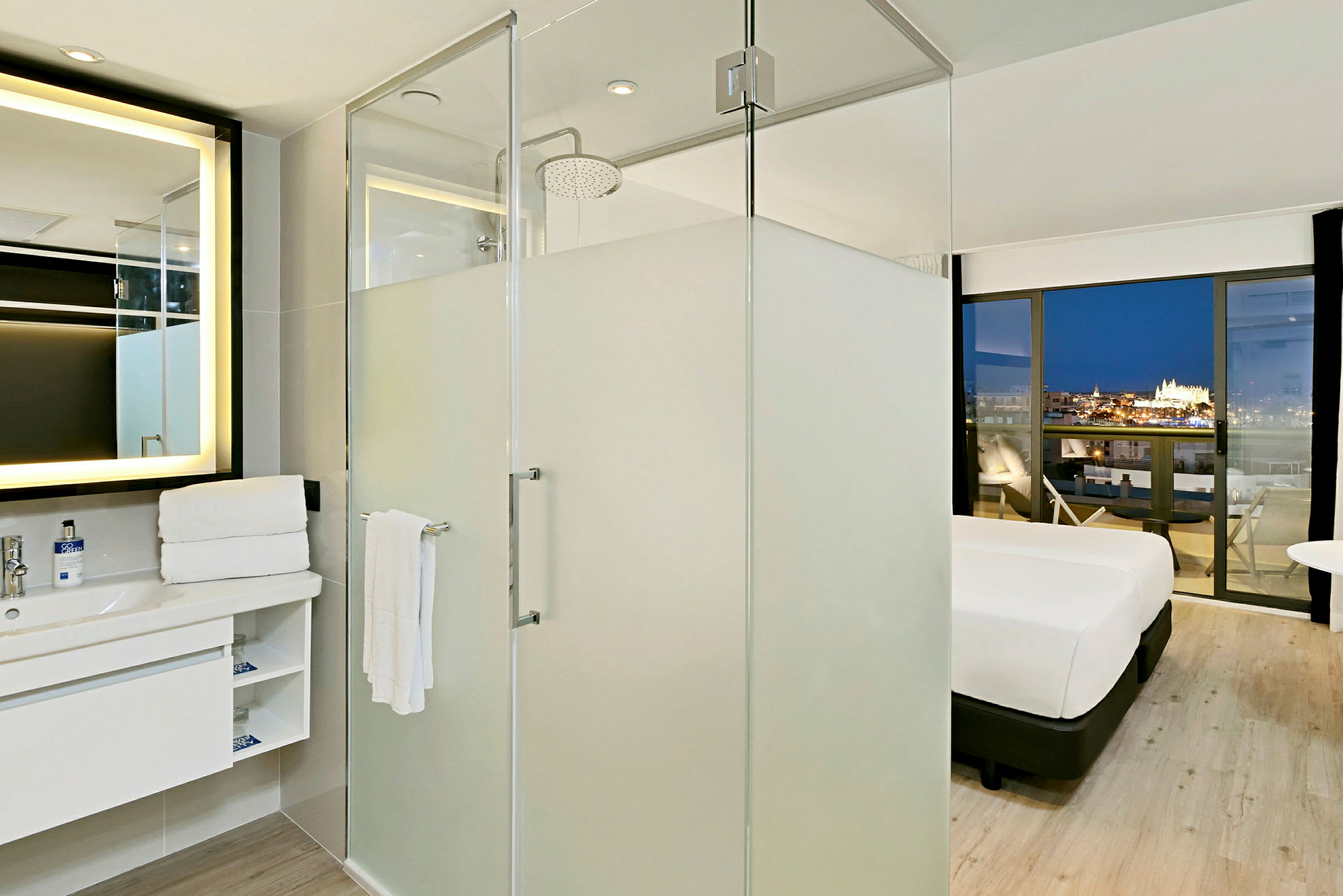 Bildet viser et moderne hotellrom med en glassvegg inn til badet, hvor dusj og servant er synlig. Rommet har en seng med utsikt mot en opplyst by gjennom store vinduer. (AI-generert tekst)