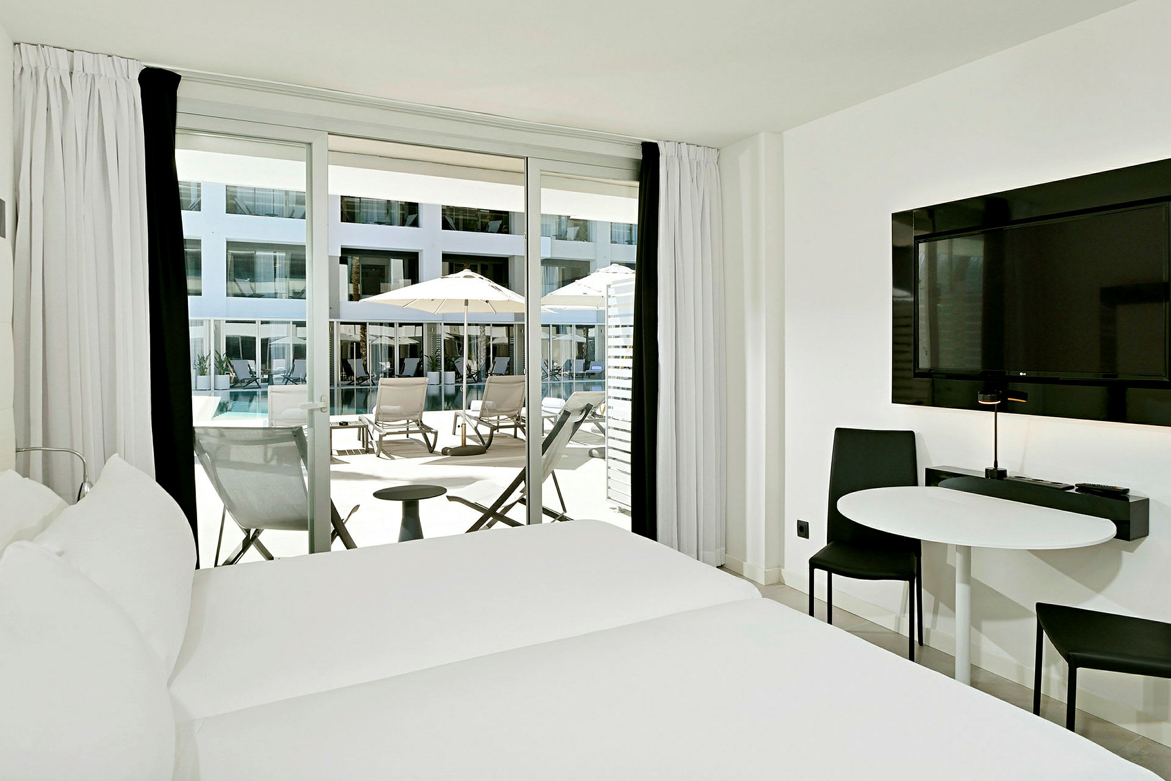 Hotellrom med seng, bord, stol og balkongdør som fører ut til terrasse med solsenger og parasoller ved et basseng. (AI-generert tekst)