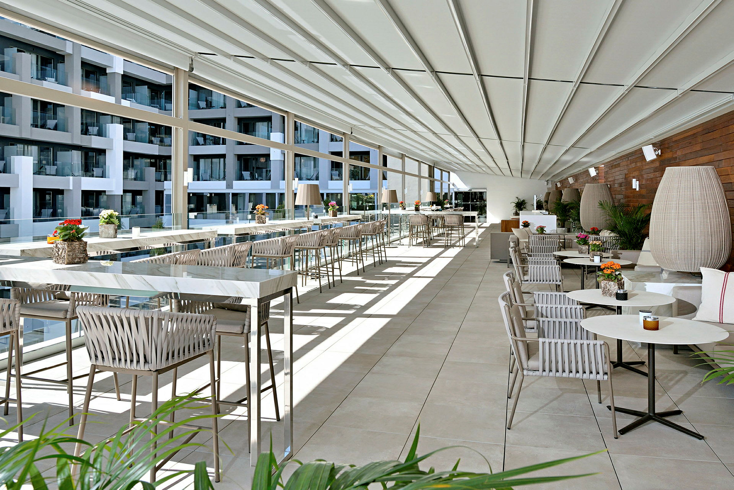 Et moderne og lyst innredet terrasse- eller restaurantområde med glassvegger, rekker av bord og stoler samt dekorative planter. (AI-generert tekst)