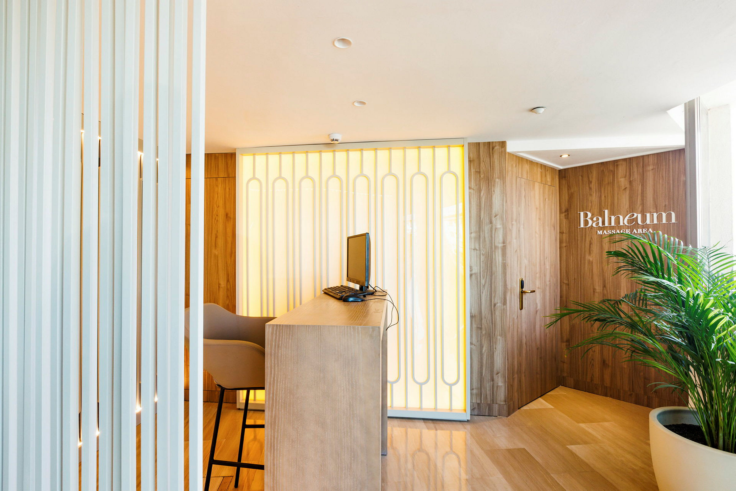 Resepsjon med bord, datamaskin og stol, med en bakgrunn av veggpaneler og en dør merket 'Balneum Massage Area'. (AI-generert tekst)