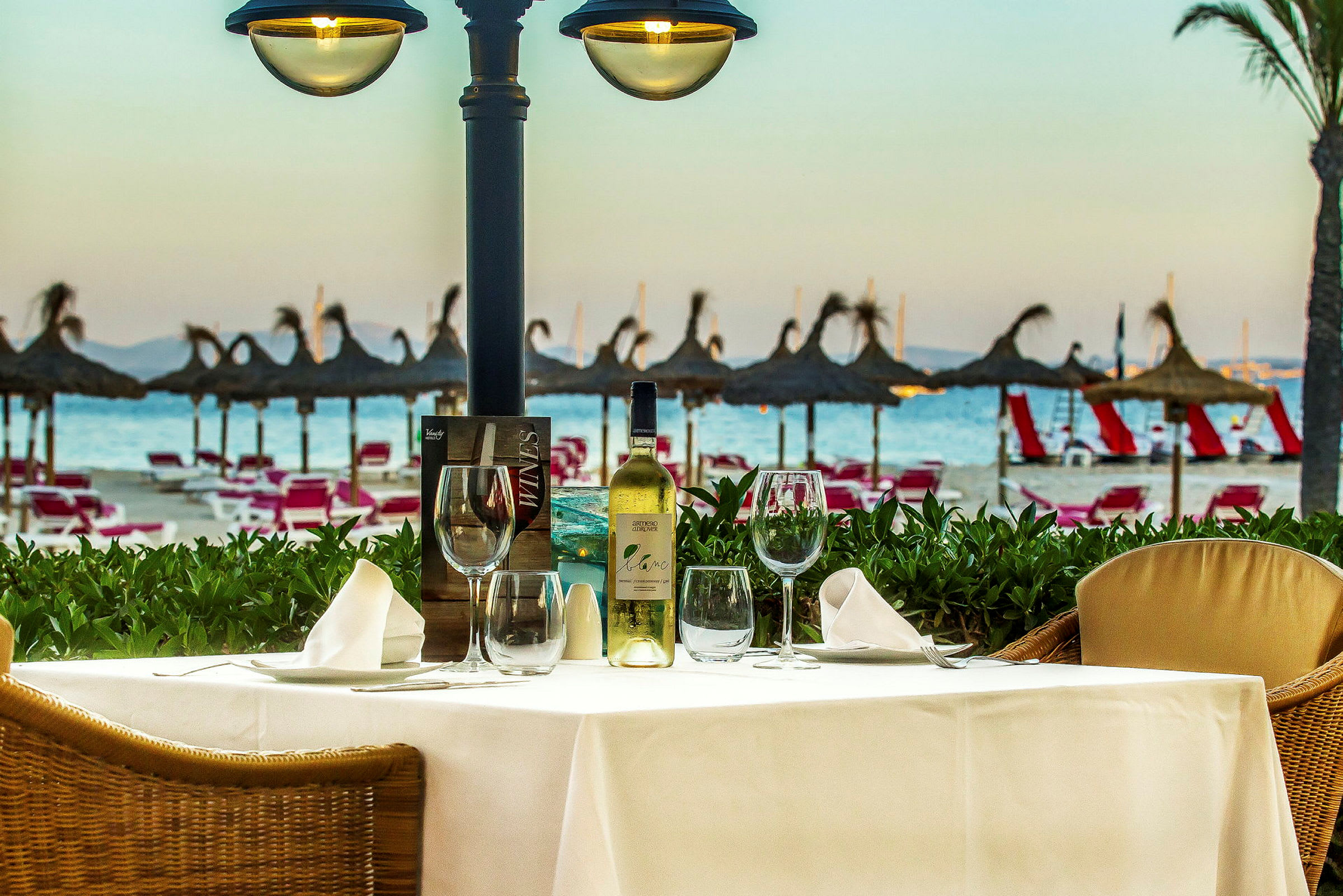 Et bord med oppdekning og en vinflaske står på en terrasse med utsikt over stranden og parasoller. (AI-generert tekst)