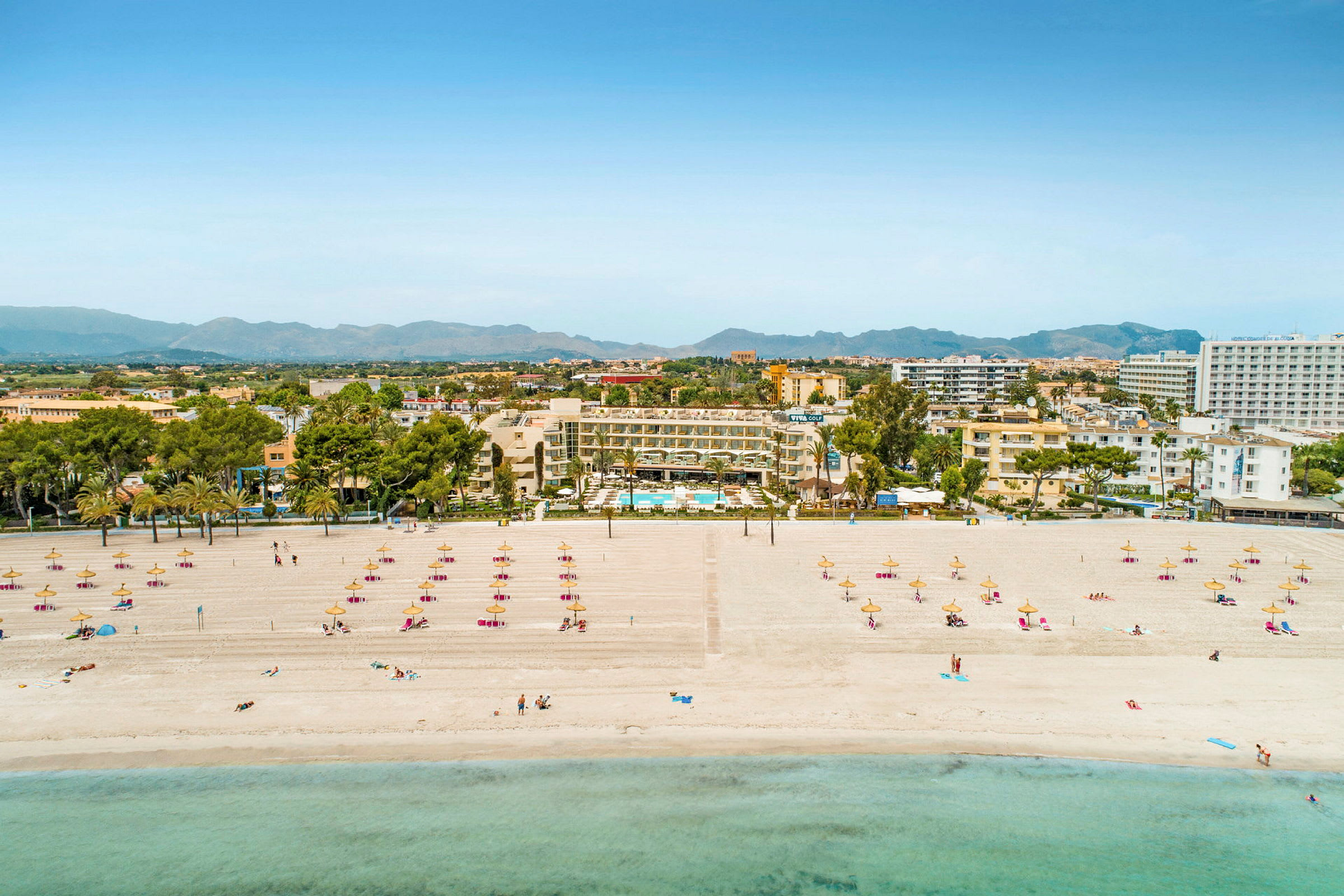 Strand med solsenger og parasoller, hotell og bysilhuett i bakgrunnen. (AI-generert tekst)