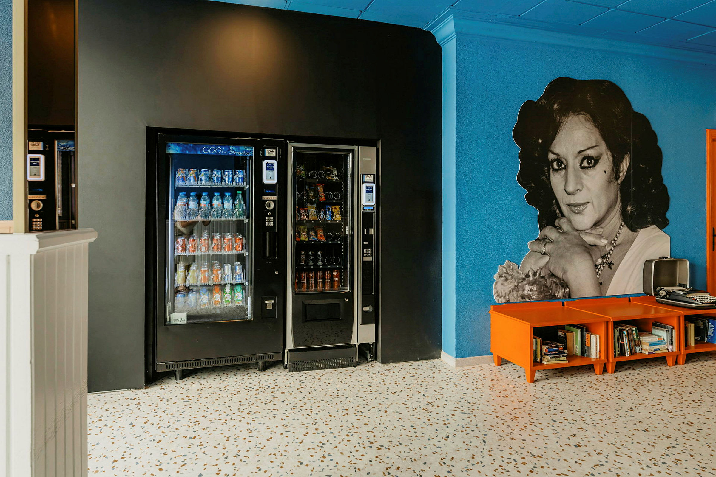 Bilde av to automater for drikker og snacks plassert mot en svart vegg, sammen med et område med oransje hyller inneholdende bøker foran en blå vegg. (AI-generert tekst)