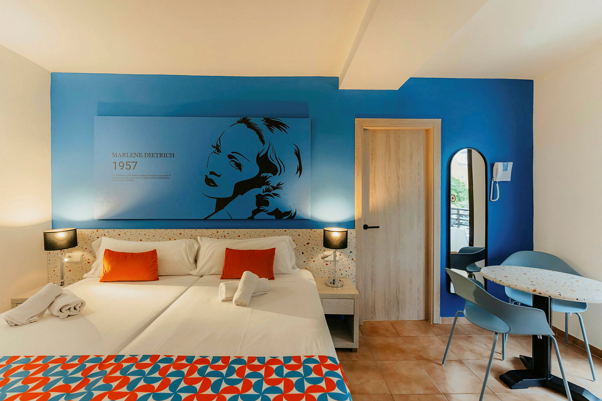 Et hotellrom med en dobbeltseng kledd i hvitt sengetøy og oransje puter. Veggen er blå og har en illustrasjon av Marlene Dietrich med tekst. Til høyre er det en dør og en liten spiseplass med et bord og to stoler. (AI-generert tekst)