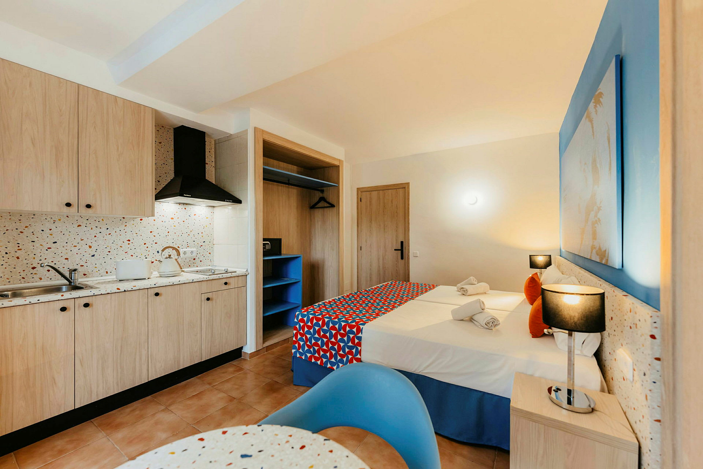 Et hotellrom med kjøkken og soveområde. Kjøkkenet har treskap, vask og komfyr. Soveområdet har to senger med fargerikt sengetøy samt nattbord og belysning. (AI-generert tekst)