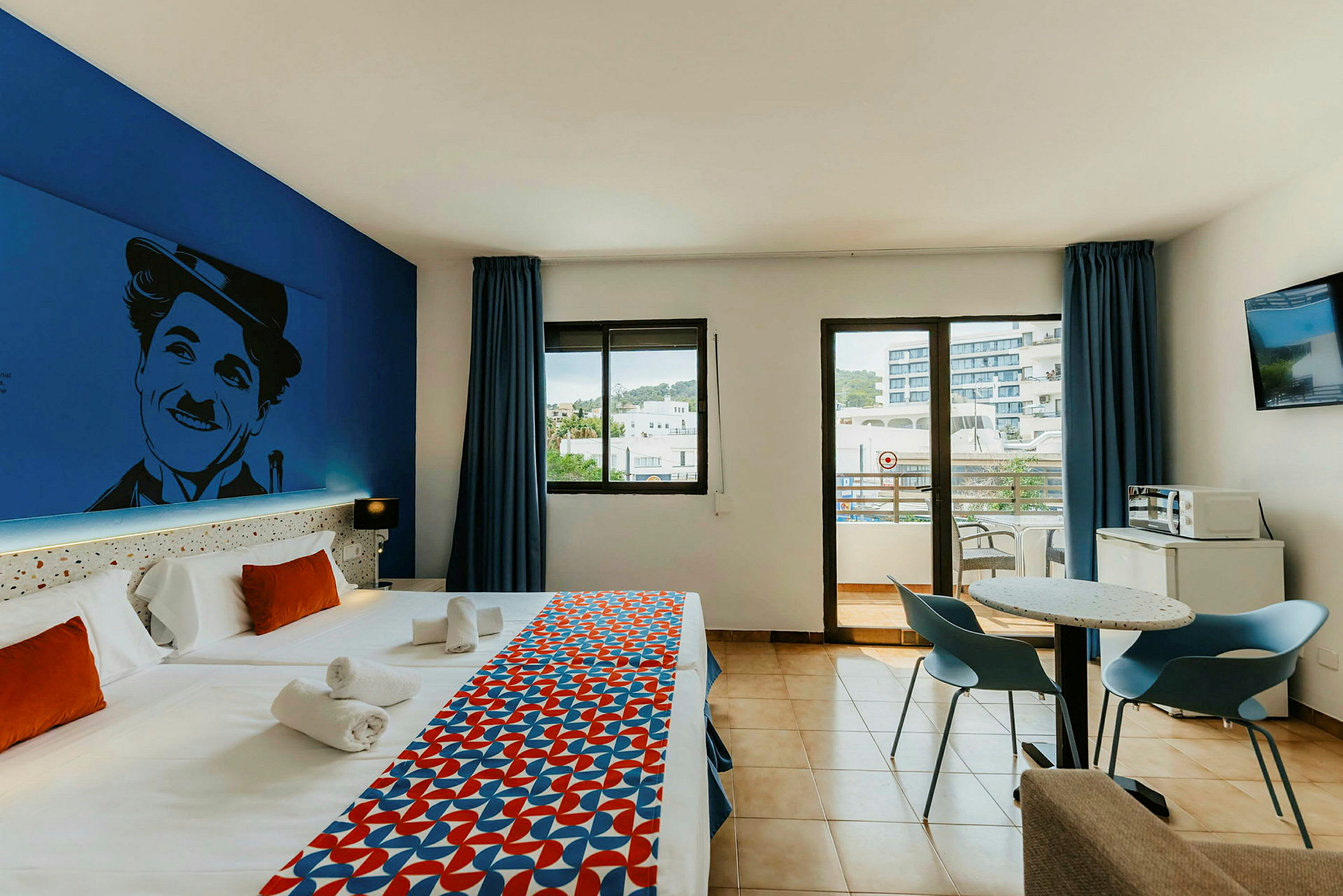 Hotellrom med to enkeltsenger, fargerik sengeteppe, veggdekorasjon, bord og stoler, samt utsikt mot en balkong. (AI-generert tekst)