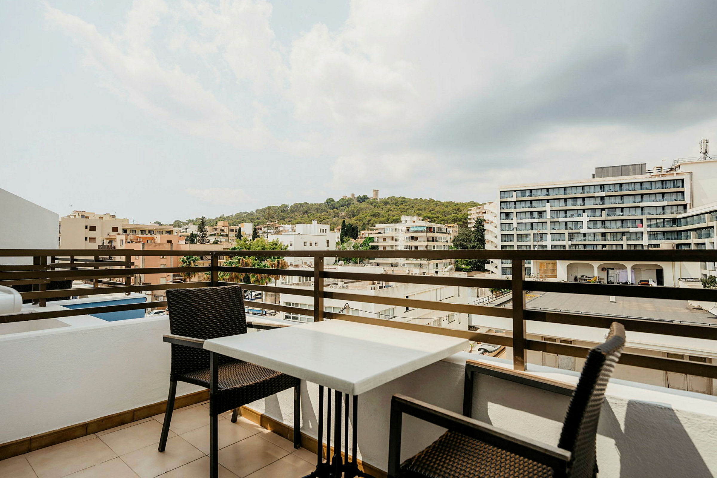 En terrasse med et bord og to stoler samt utsikt over byen med bygninger og grønne åser i bakgrunnen. (AI-generert tekst)