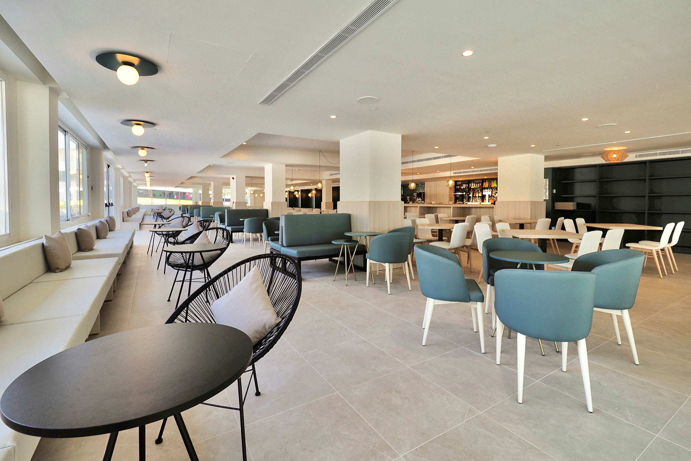 Bilde av en moderne og romslig innendørs lounge eller restaurant, med bord og stoler, samt store vinduer som slipper inn dagslys. (AI-generert tekst)