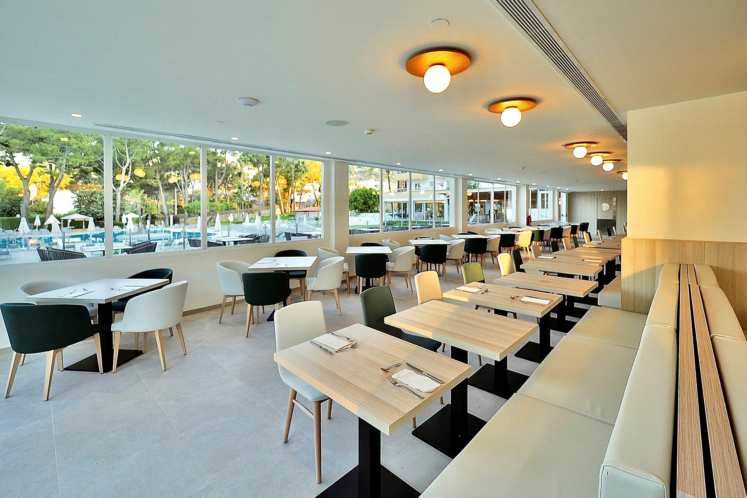En moderne restaurant med vinduer som vender mot et utendørs basseng og grøntområder. Bord og stoler er lyse og enkelt designet. (AI-generert tekst)