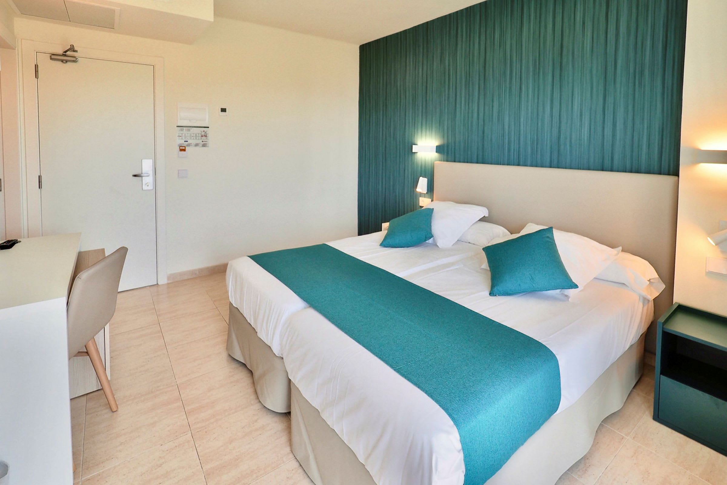 Et hotellrom med en dobbeltseng med blå dekorative puter og sengeteppe. En lampe og et nattbord vises ved siden av sengen. (AI-generert tekst)