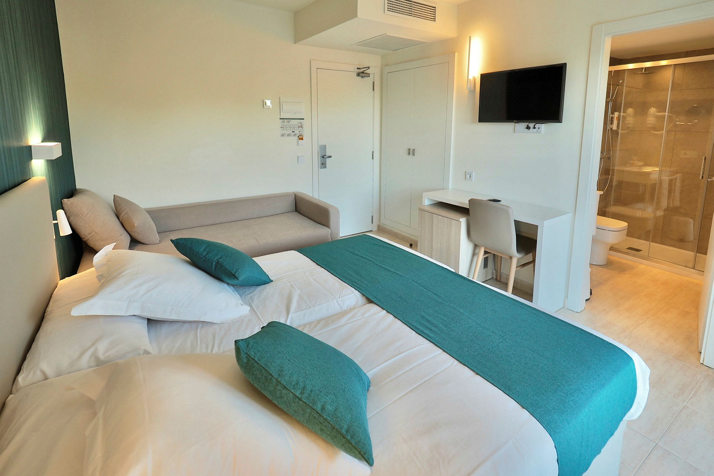 Et hotellrom med en seng med hvite laken og dekorative grønne puter samt et matchende grønt sengeteppe. På rommet er det en lys sofa, et skrivebord med stol og en TV på veggen. Badet med dusj kan ses gjennom en glassdør til høyre. (AI-generert tekst)