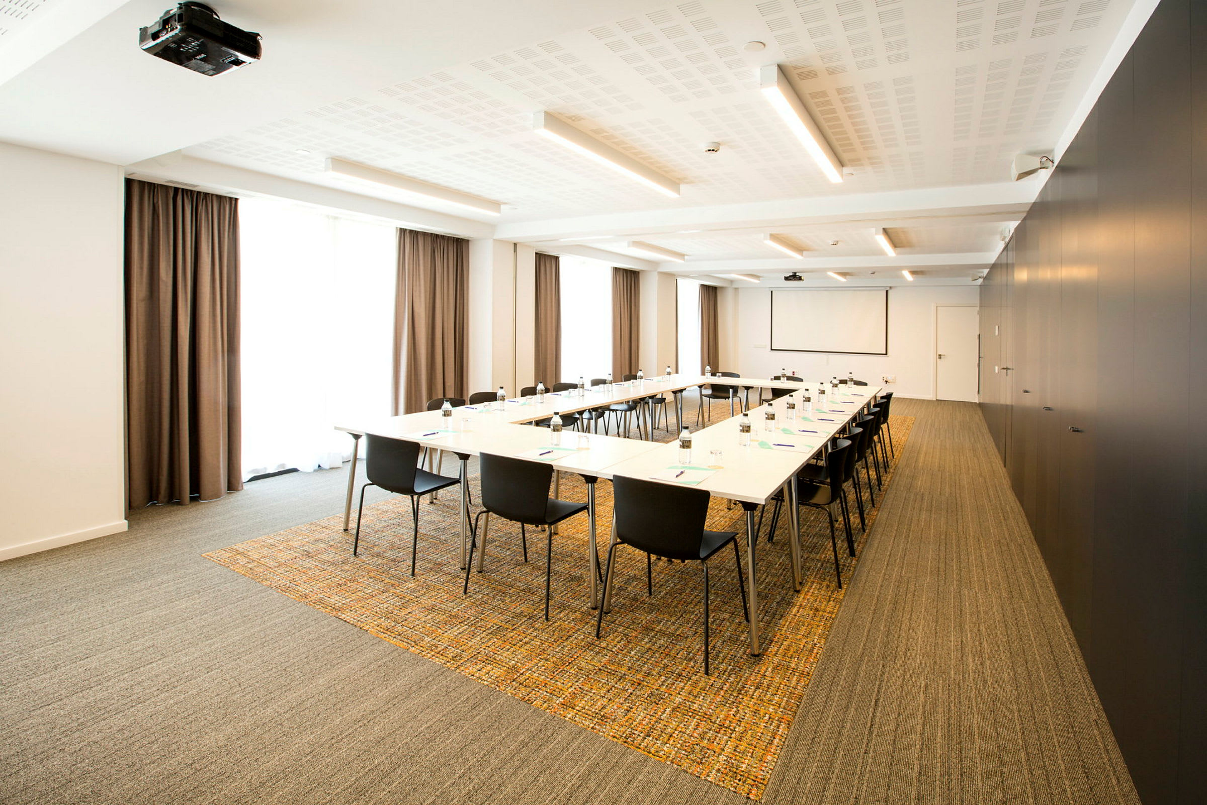 Konferanserom med bord arrangert i U-form, omgitt av svarte stoler. Vinduer med gardiner er synlige i bakgrunnen. (AI-generert tekst)