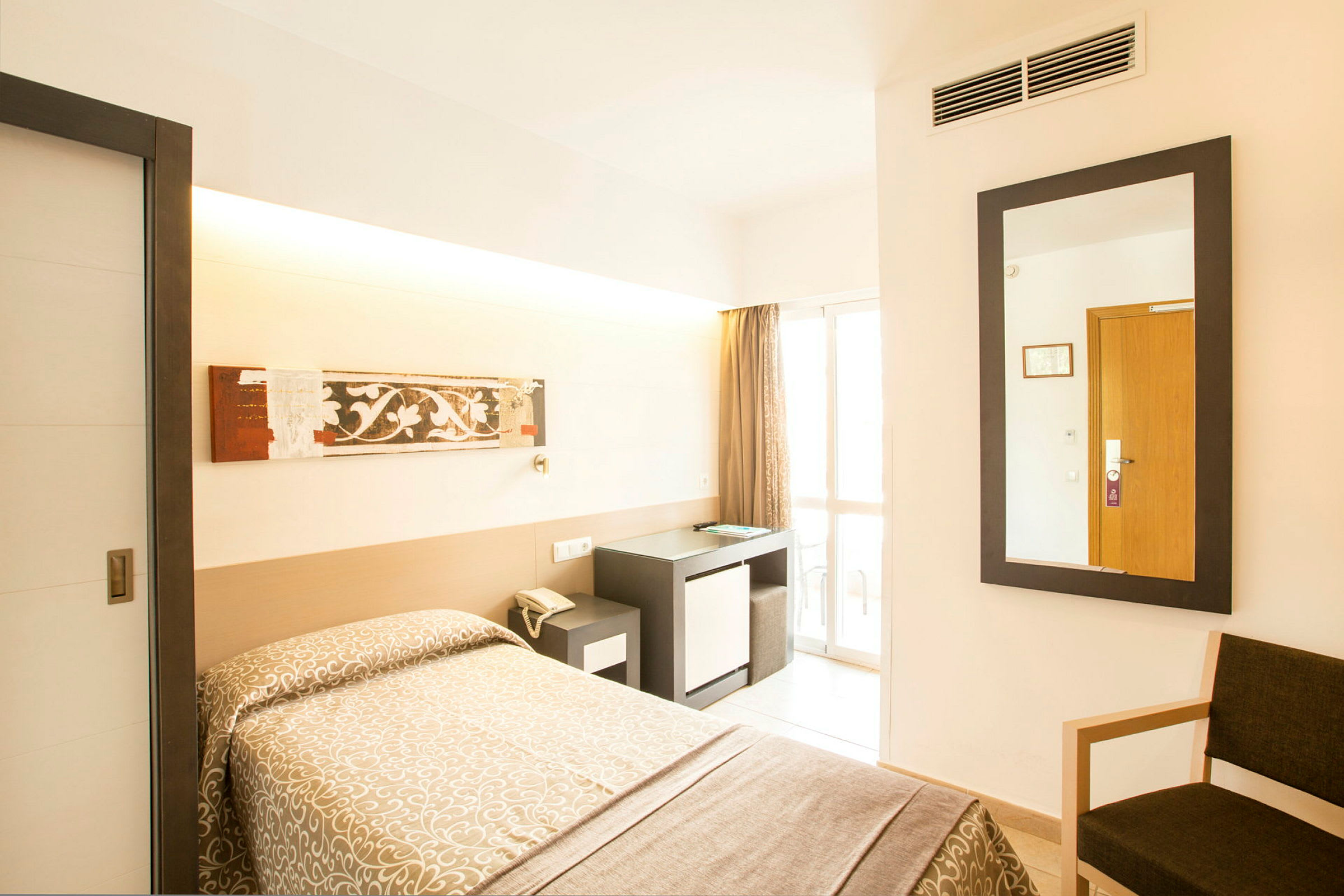 Hotellrom med en enkeltseng dekket med et mønstret sengeteppe, et skrivebord med en stol og et speil på veggen. En glassdør fører ut til en balkong. (AI-generert tekst)